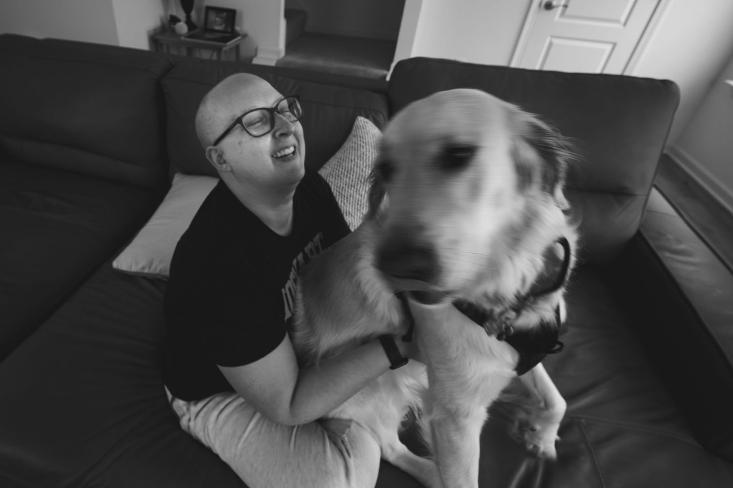 Bald man with glasses smiling while petting a golden retriever dog on a gray couch in black and white photo.