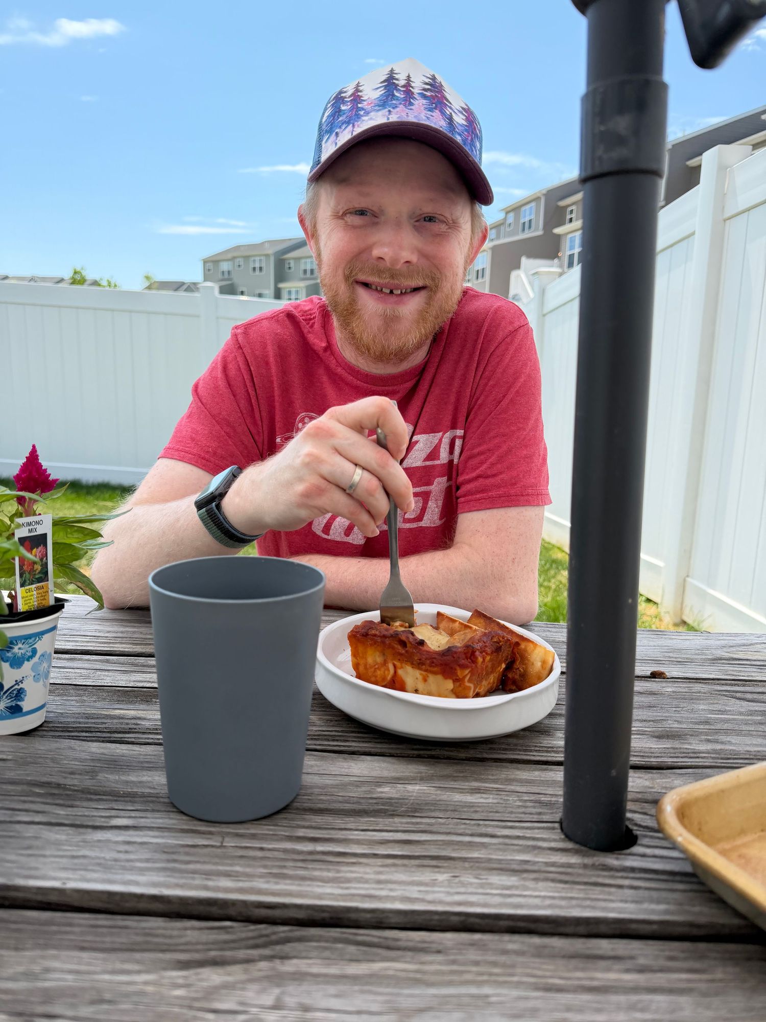 Smiling man in red shirt and cap eating BBQ food at outdoor picnic table with gray cup on sunny day.