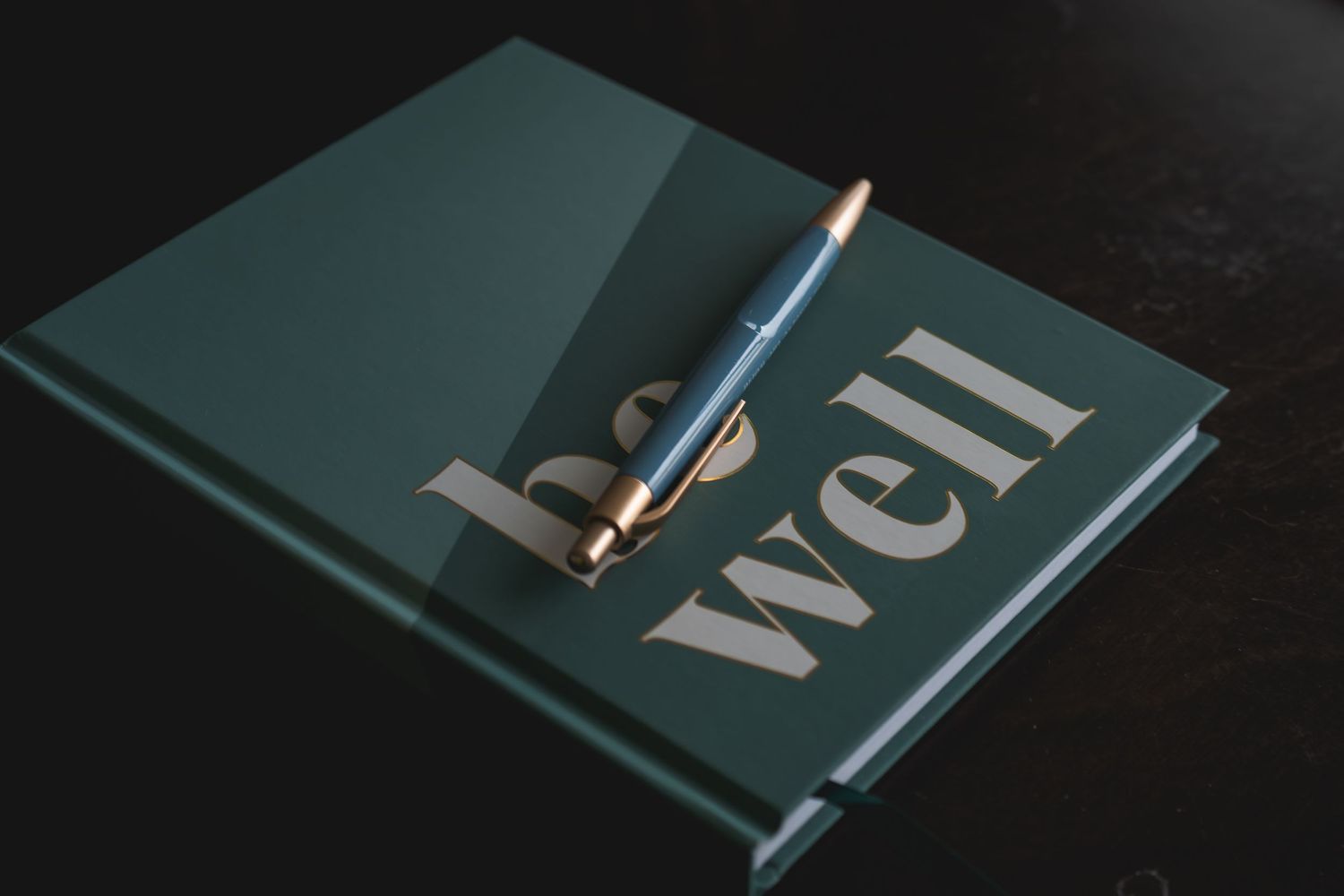 A teal hardcover journal titled 'be well' with a rose gold pen resting on top, placed on a dark surface.
