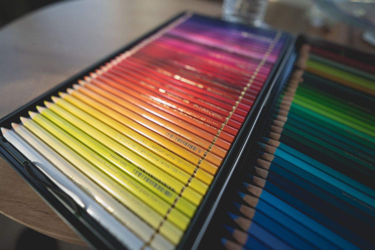 A large set of colorful pencils arranged in rainbow gradient order in a black case on a wooden table.