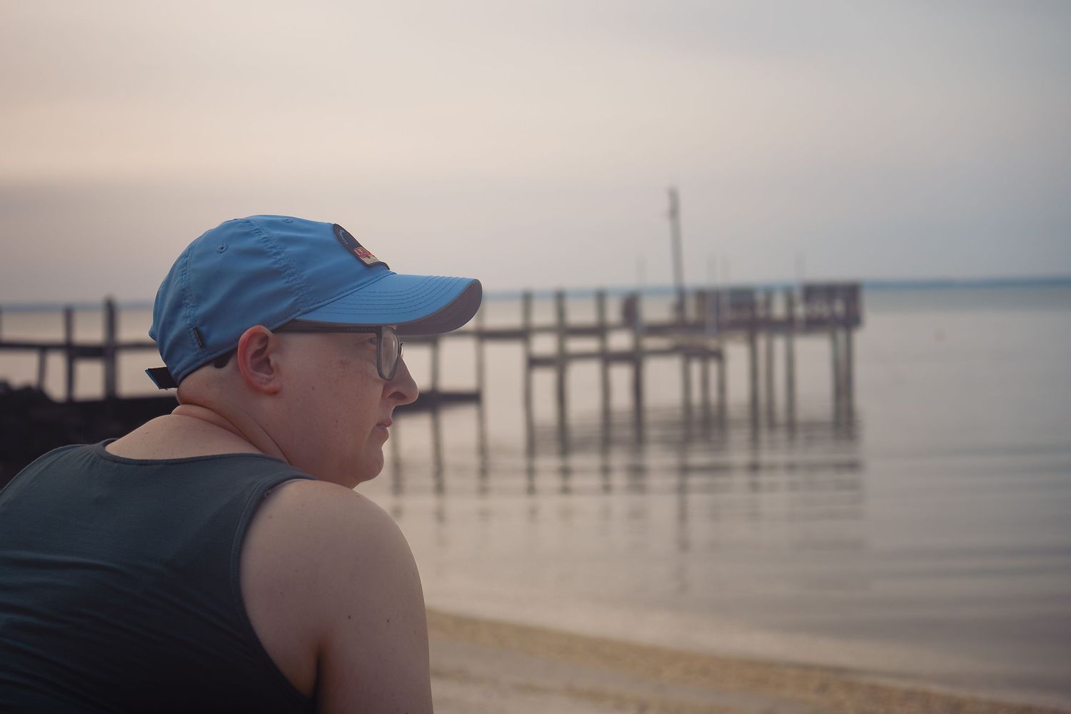Person in blue cap and tank top gazing at a calm waterway with a wooden pier at dusk.