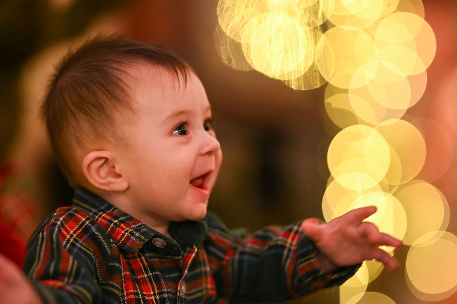 A baby in a plaid shirt looks up with wonder at twinkling holiday lights creating a magical bokeh effect in the background.