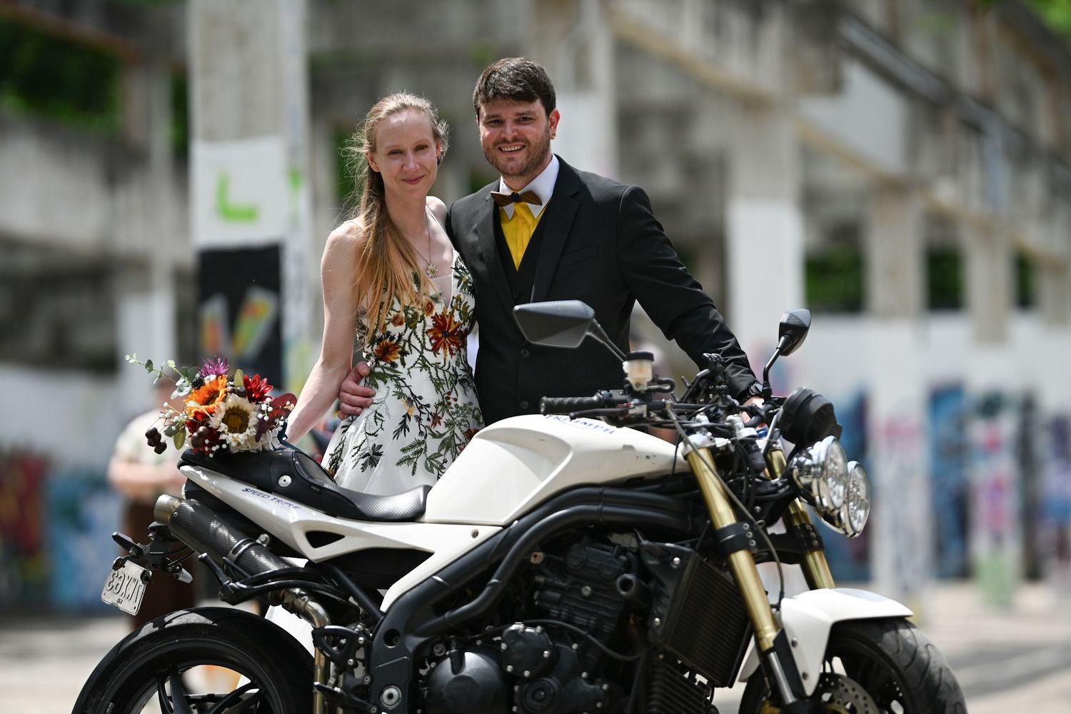 A couple in formal wedding attire poses on a white motorcycle in an urban setting with graffiti walls.