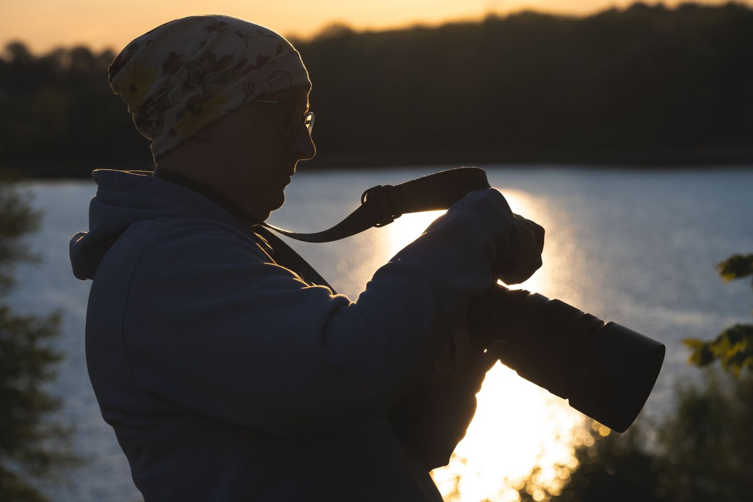 Photographer silhouetted against golden sunset by a lake, holding a camera with telephoto lens outdoors.