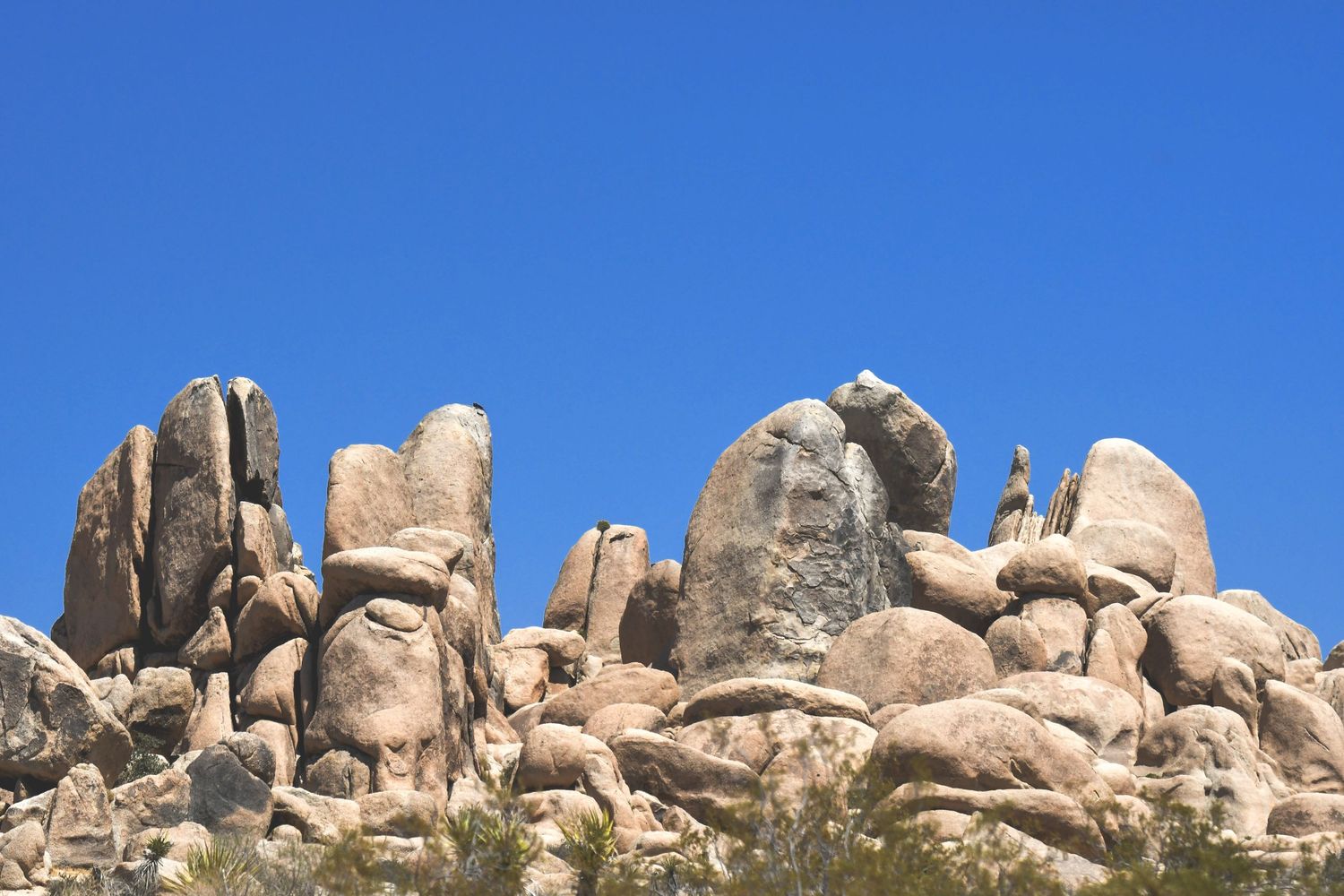 Large granite boulders stacked against a clear blue sky in a desert landscape with small shrubs below.