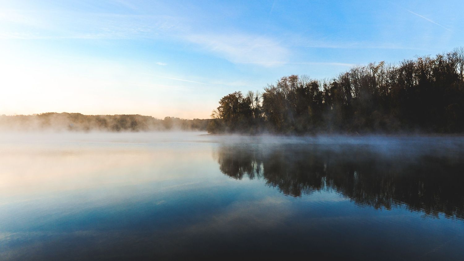 Misty morning lake with calm reflective water, surrounded by autumn trees under a soft blue sky at sunrise.