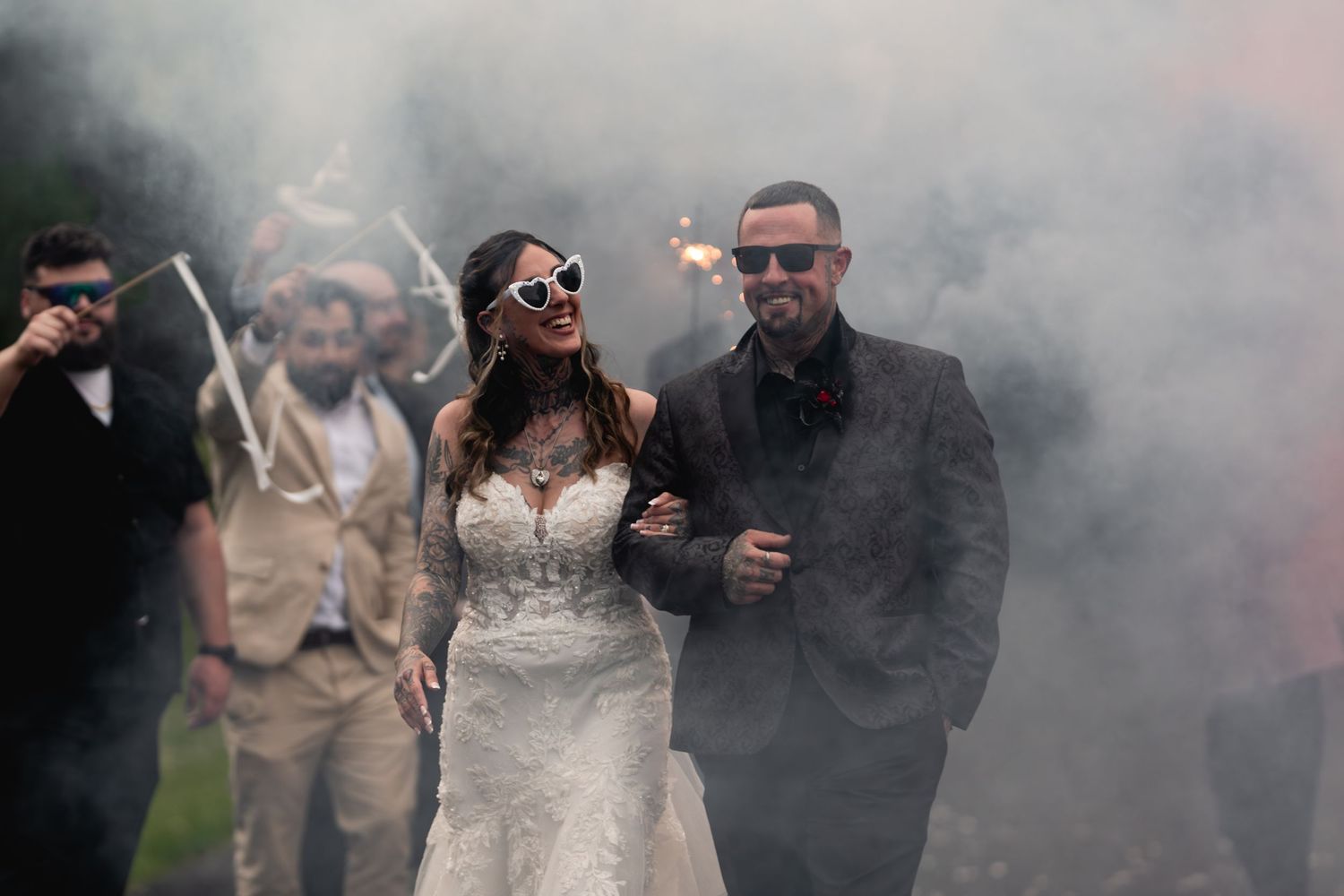 Bride and groom walk through dramatic smoke surrounded by guests holding sparklers at outdoor wedding celebration.