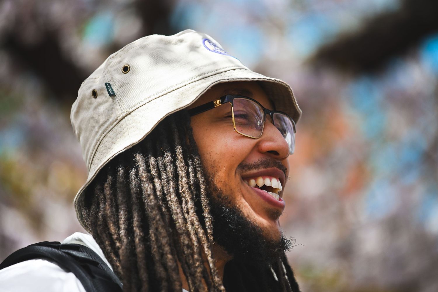 Individual with dreadlocks and glasses laughs joyfully while wearing a bucket hat outdoors.