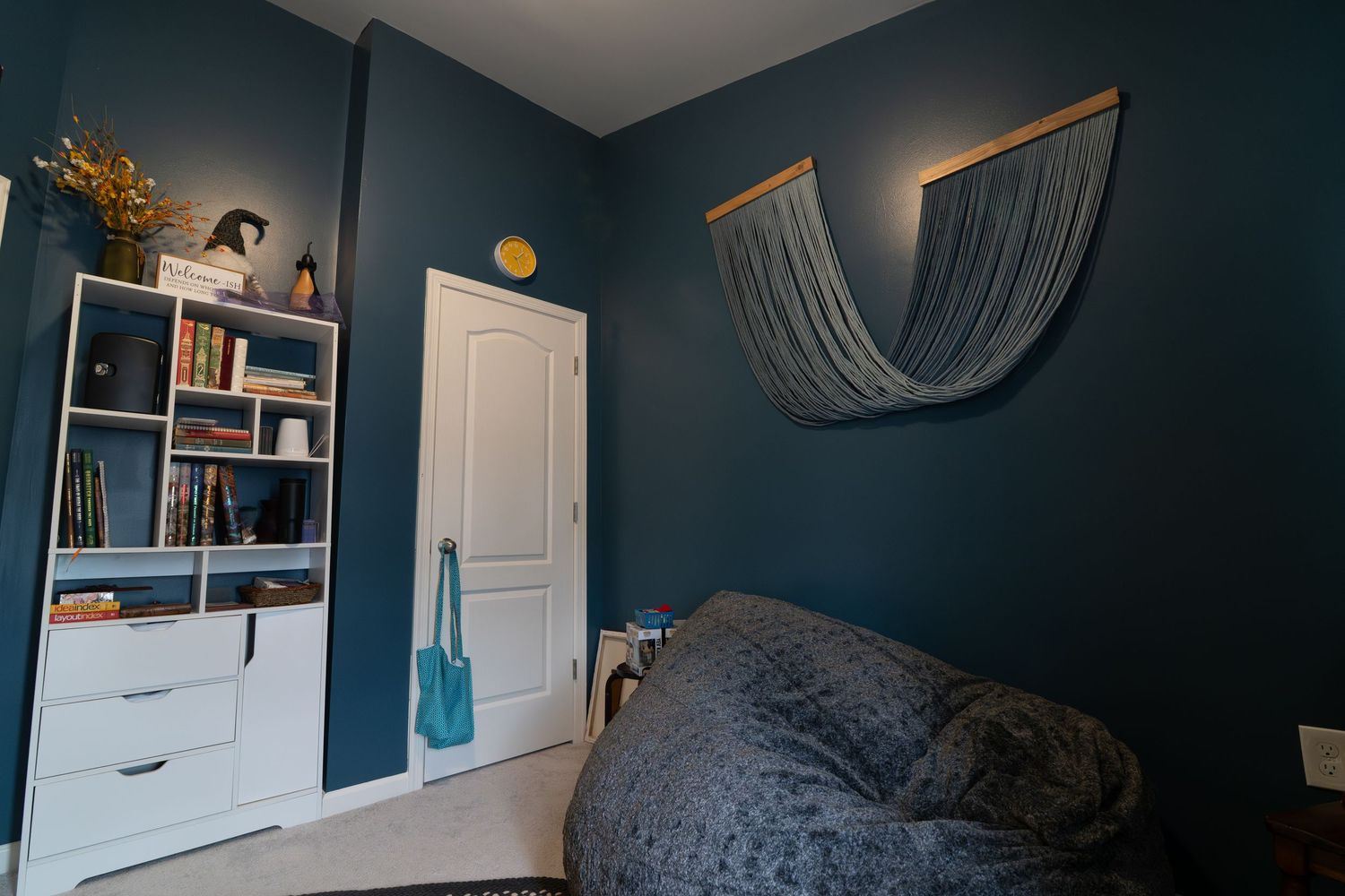 Cozy bedroom with teal blue walls, white bookshelf, bean bag chair, and decorative fabric wall art.