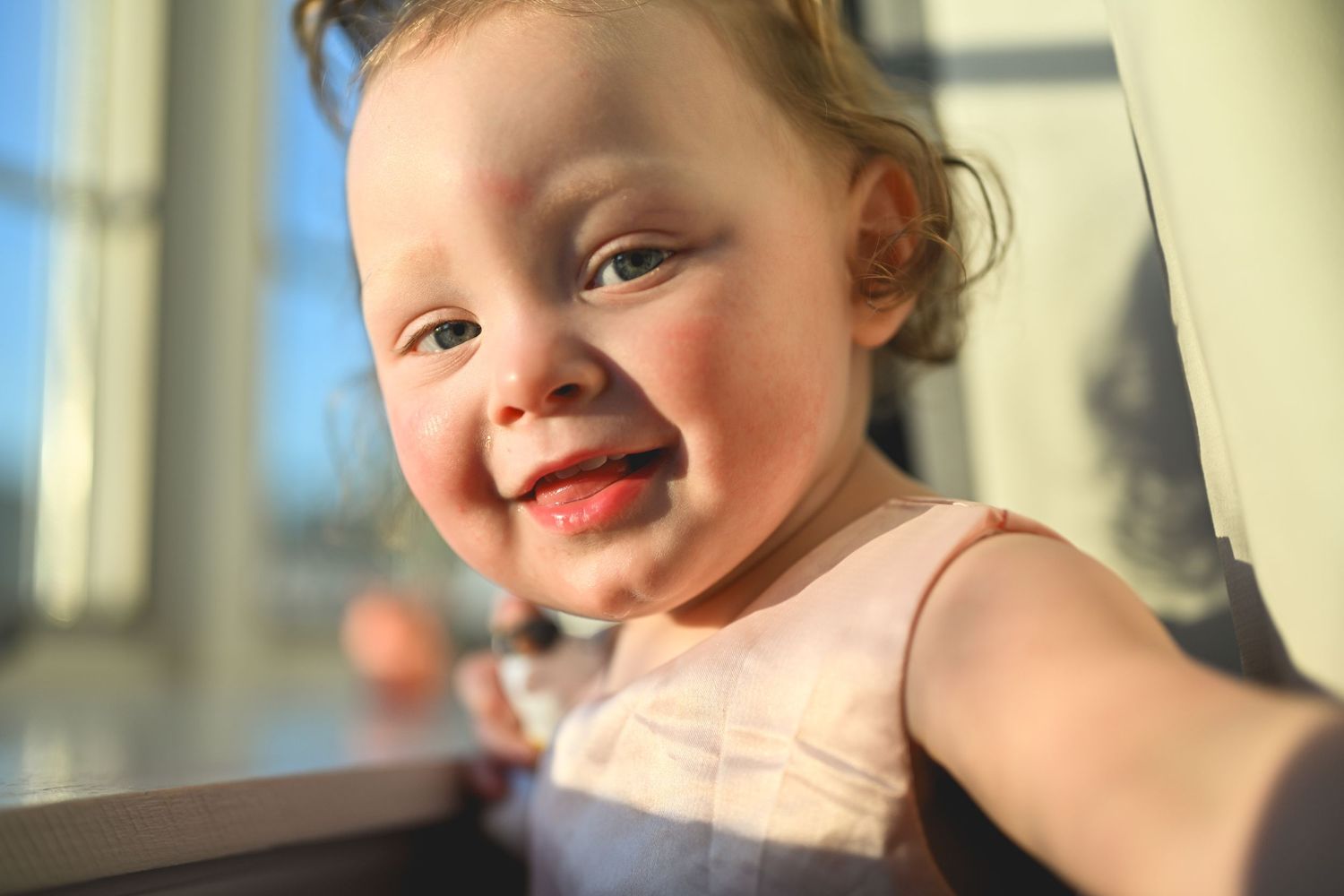 A young child wearing a sleeveless dress smiles brightly while sitting indoors in warm lighting.
