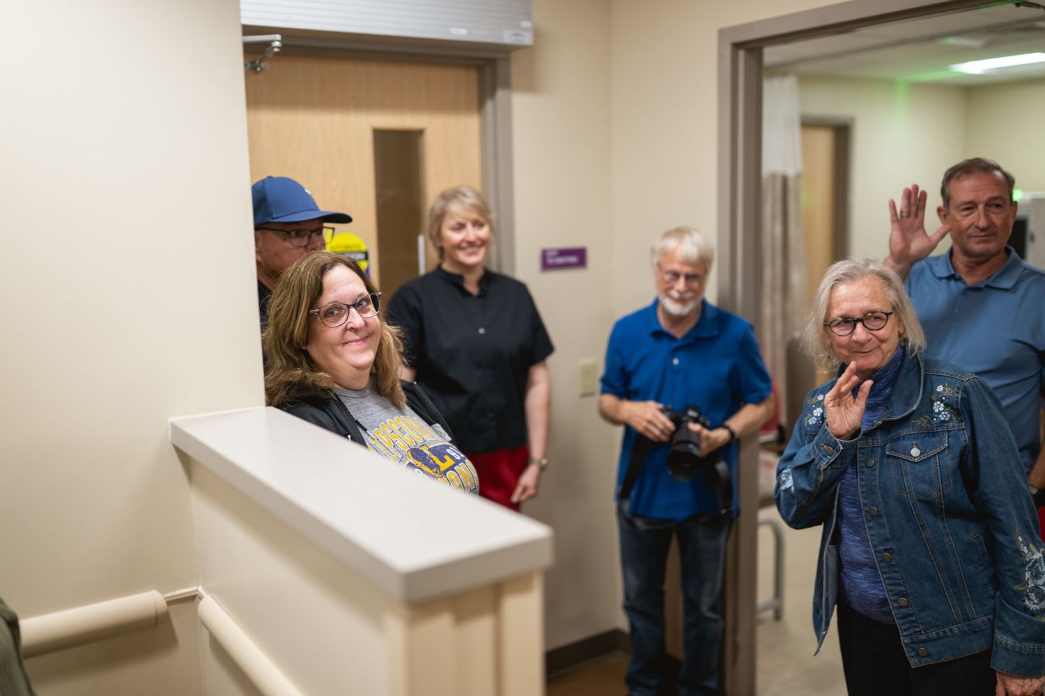 Group of adults smiling and waving in a hallway, gathered for a casual indoor event or facility tour.