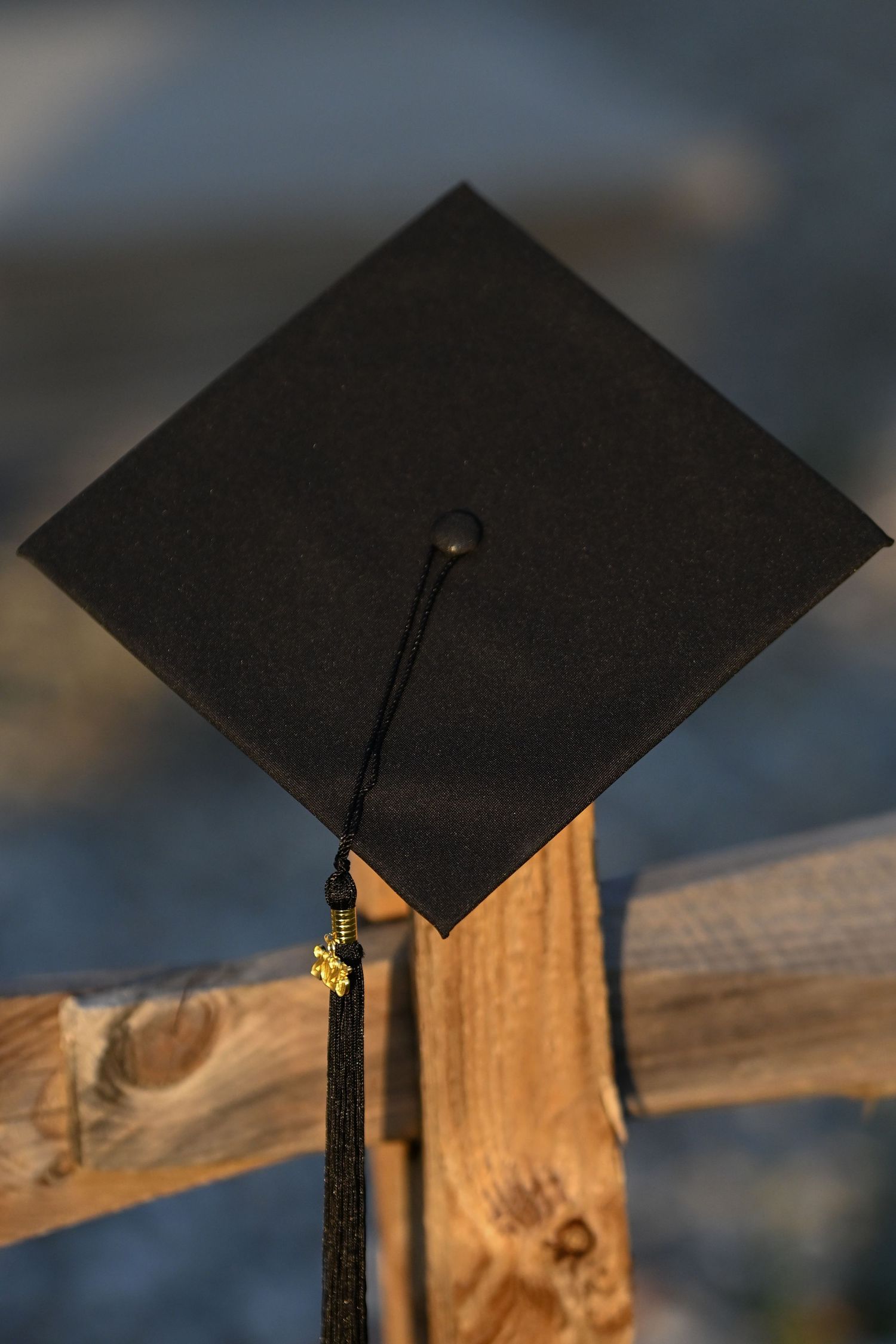 A black graduation cap with hanging tassel rests on a wooden railing against a blurred background.