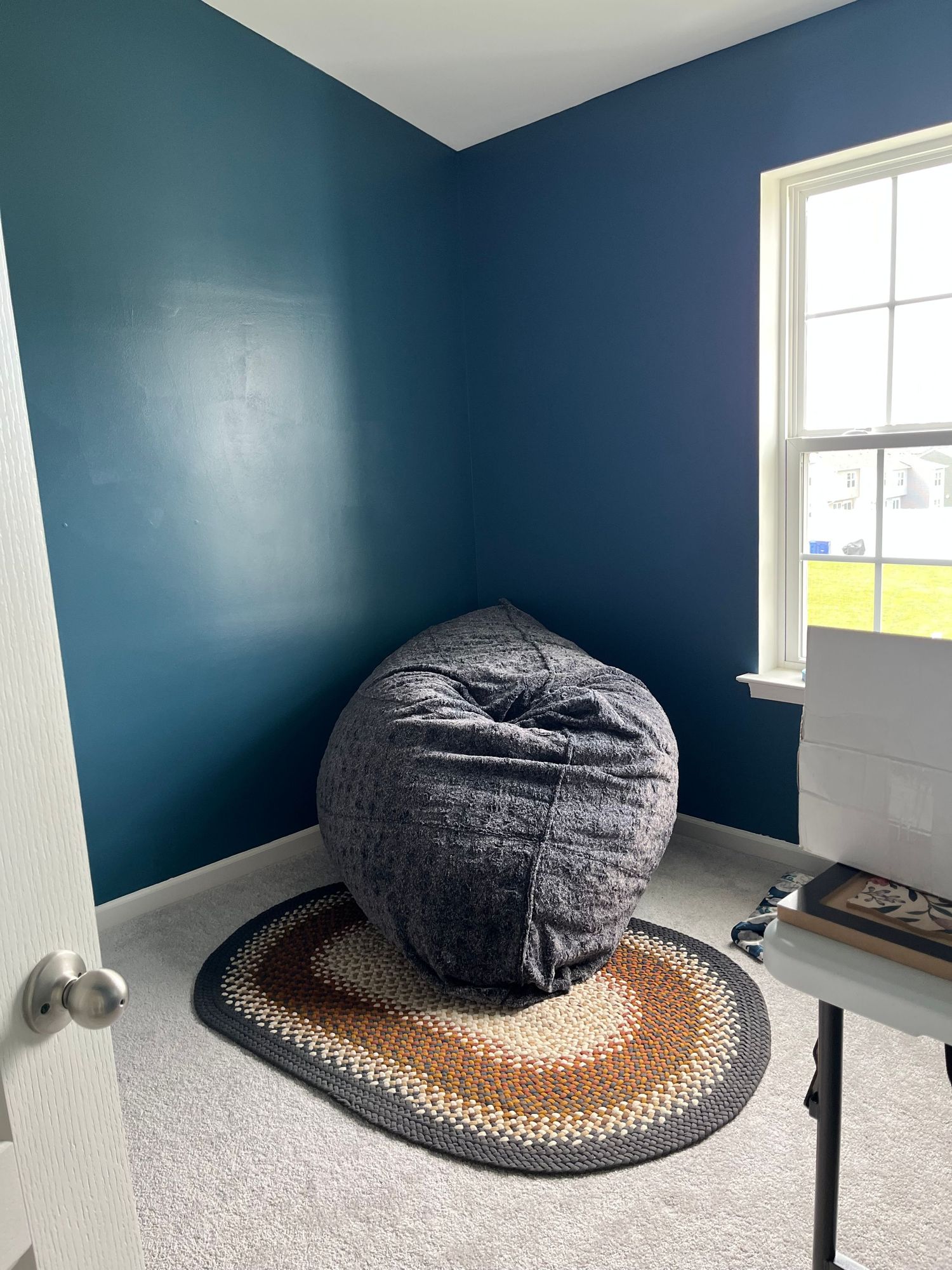 A cozy corner with a gray bean bag chair on a braided rug against dark teal walls near a bright window.