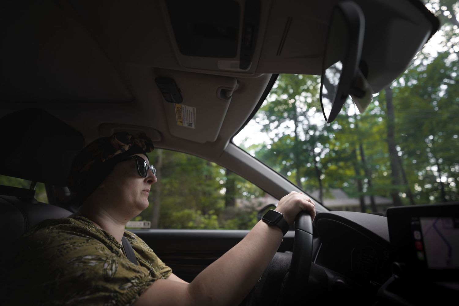 Person wearing sunglasses and camo hat driving a car through a tree-lined road during daytime.