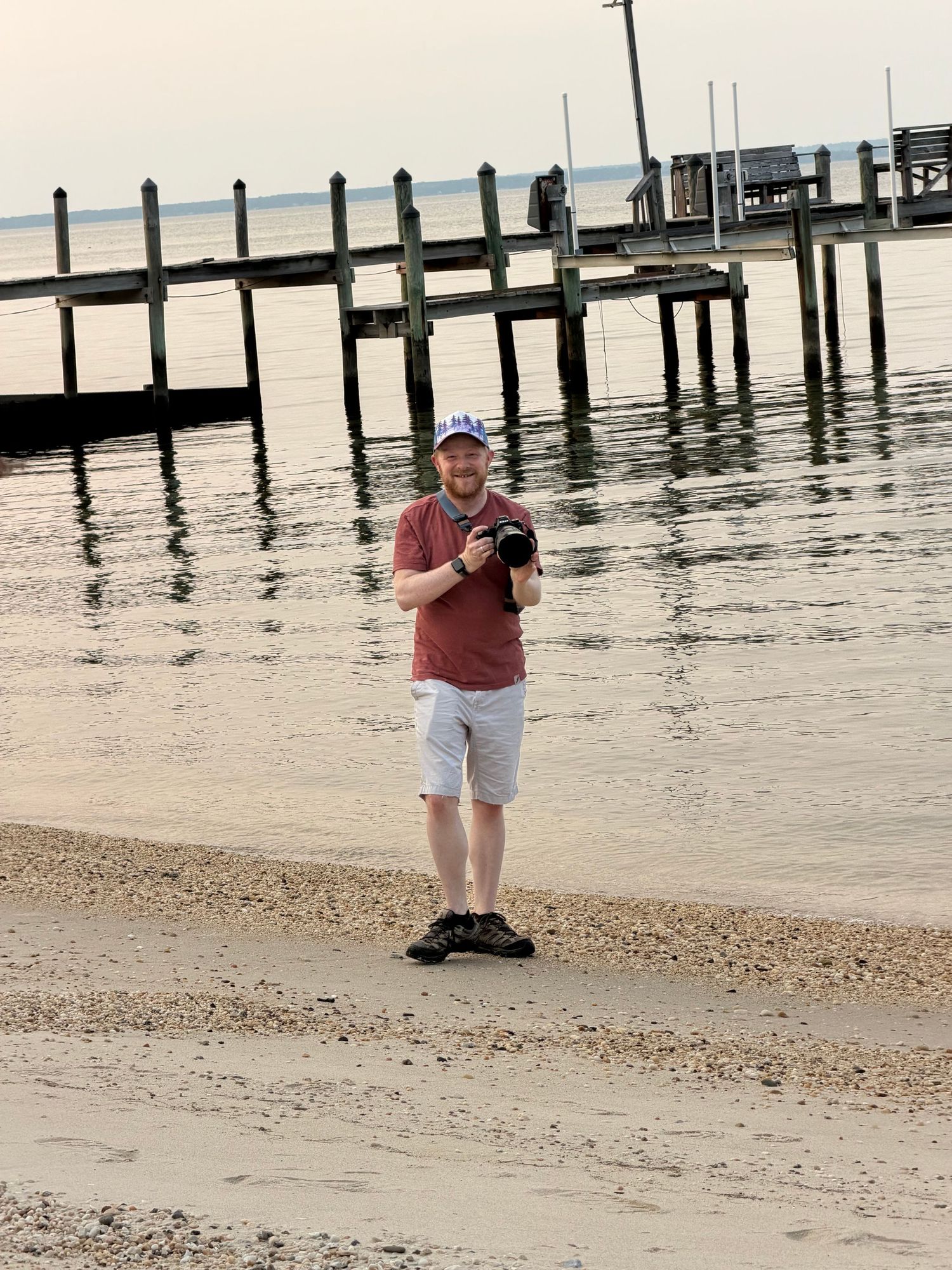 Man holding camera on sandy beach near wooden pier on calm water on overcast day.