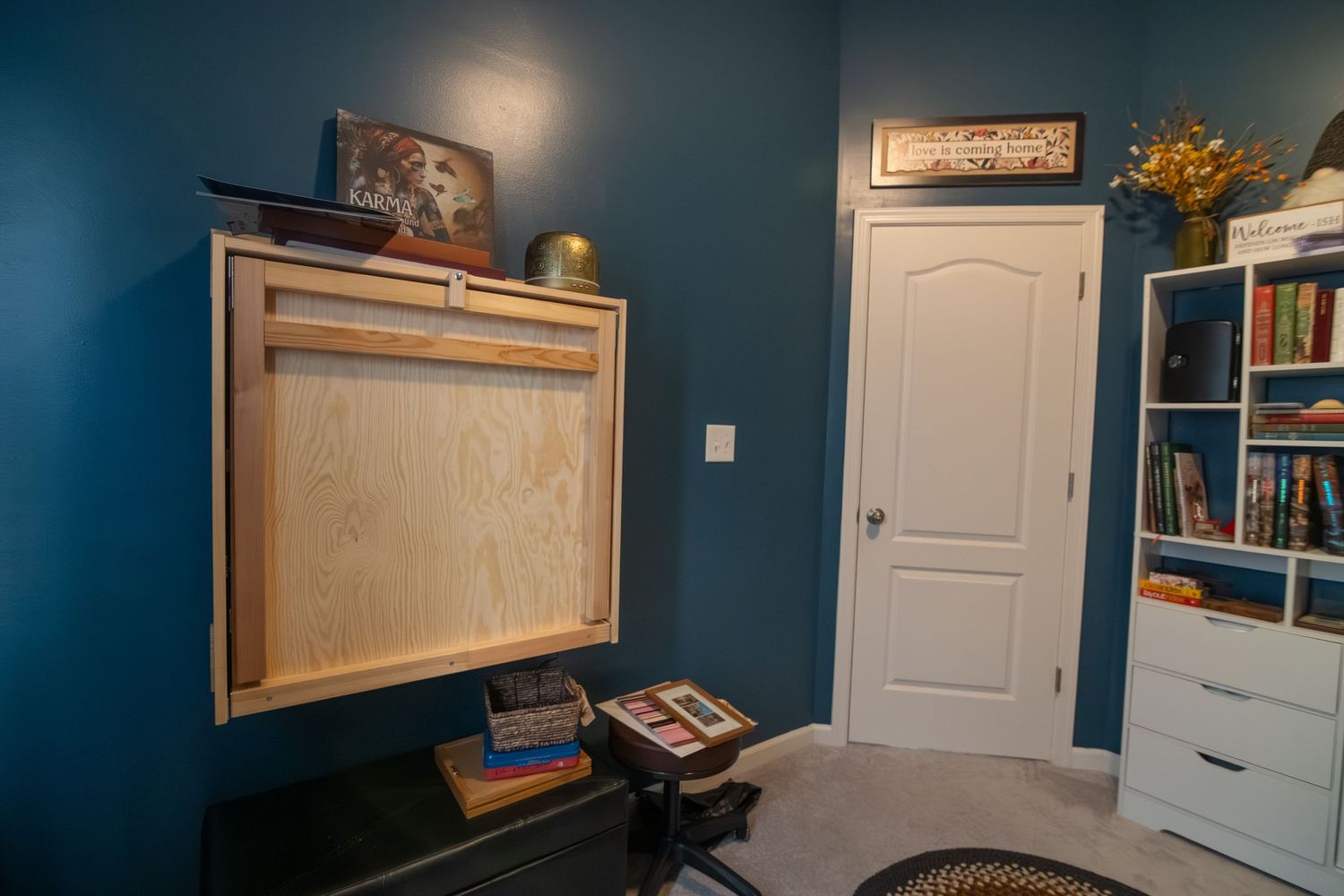 Wall-mounted fold-down desk in a teal blue home office with white door, bookshelf, and carpet flooring.