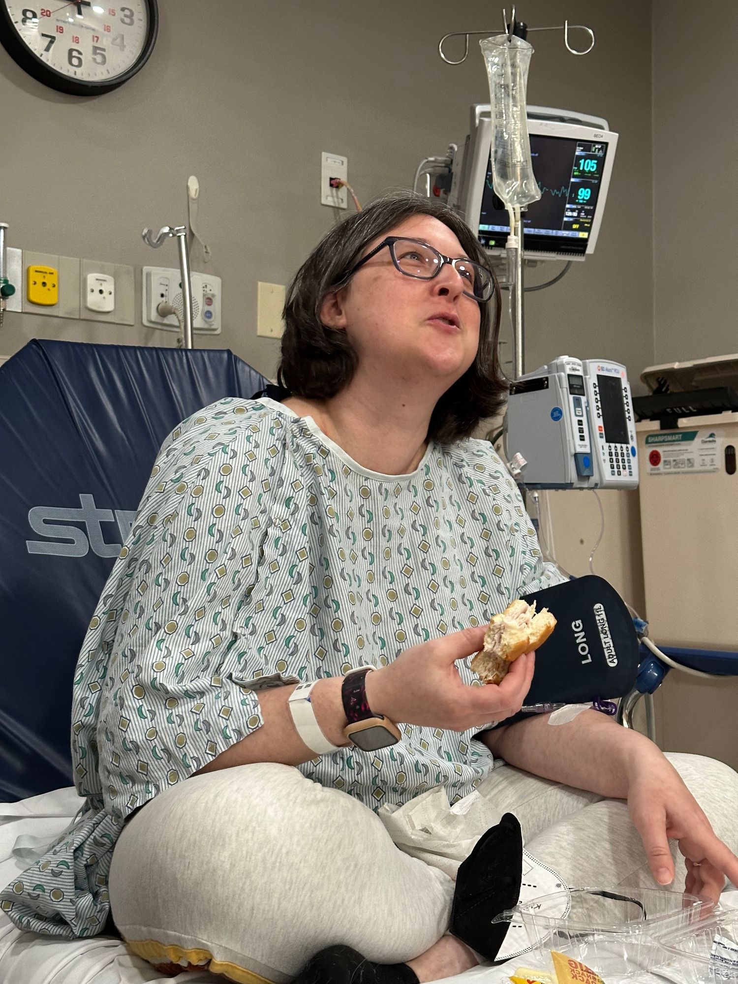 A patient in a hospital gown sits cross-legged on a hospital bed, eating food while connected to medical equipment.