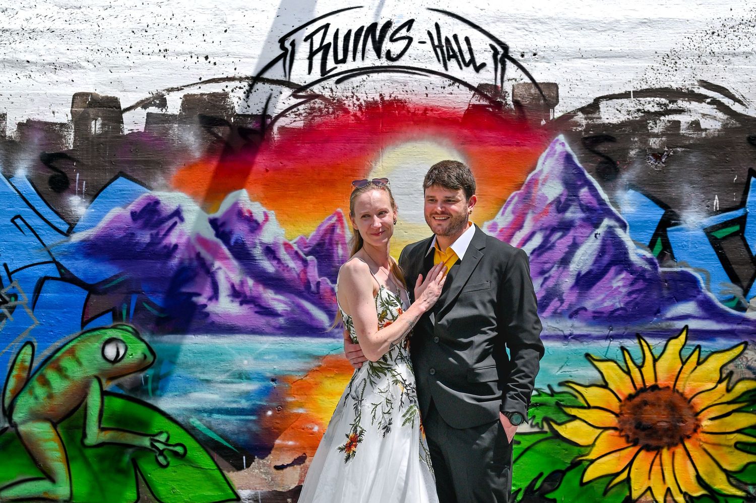 A couple poses in wedding attire in front of a vibrant graffiti mural featuring mountains, a sunflower and a green frog.