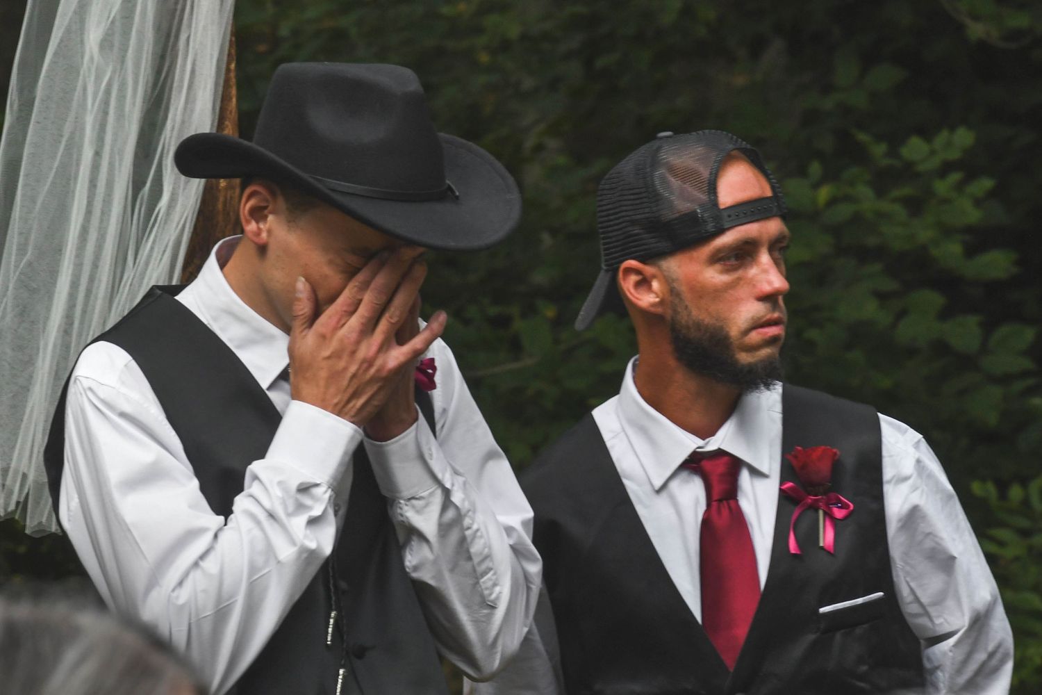 Two people wearing vests and formal attire stand together at an emotional outdoor wedding ceremony.