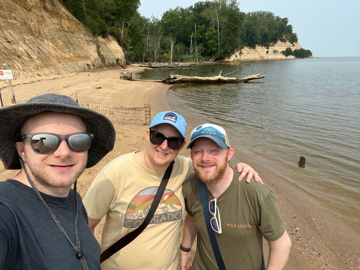 Three friends smiling for a selfie on a sandy beach with rocky cliffs and calm water in the background.
