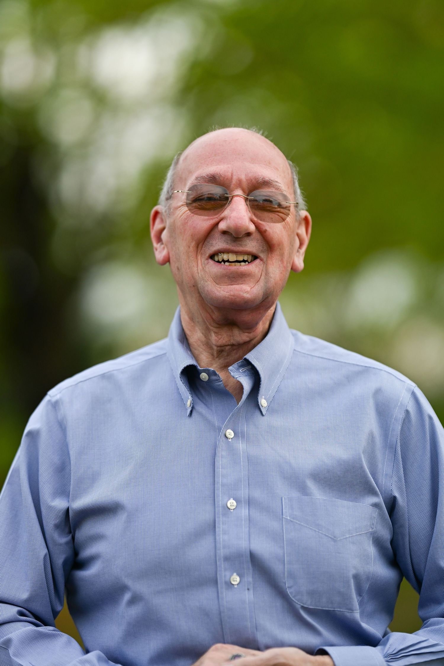 Portrait of someone wearing a light blue button-down shirt and glasses smiling outdoors with soft green foliage in background.