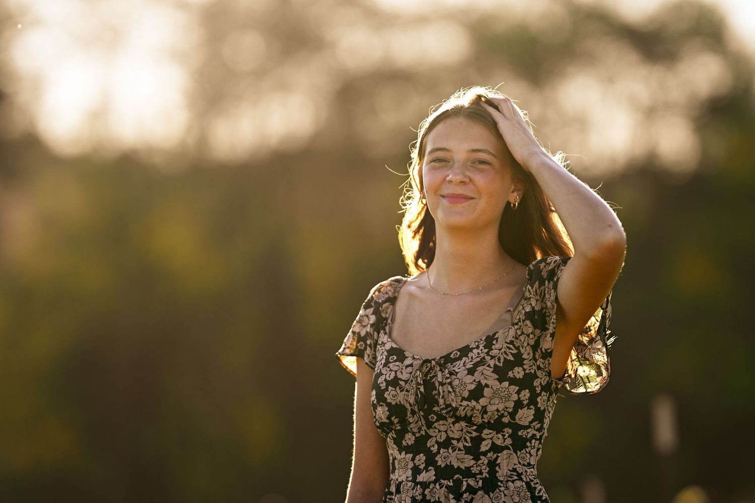 Young woman in floral dress smiling outdoors in golden sunlight, hand in hair, with blurred trees in background.