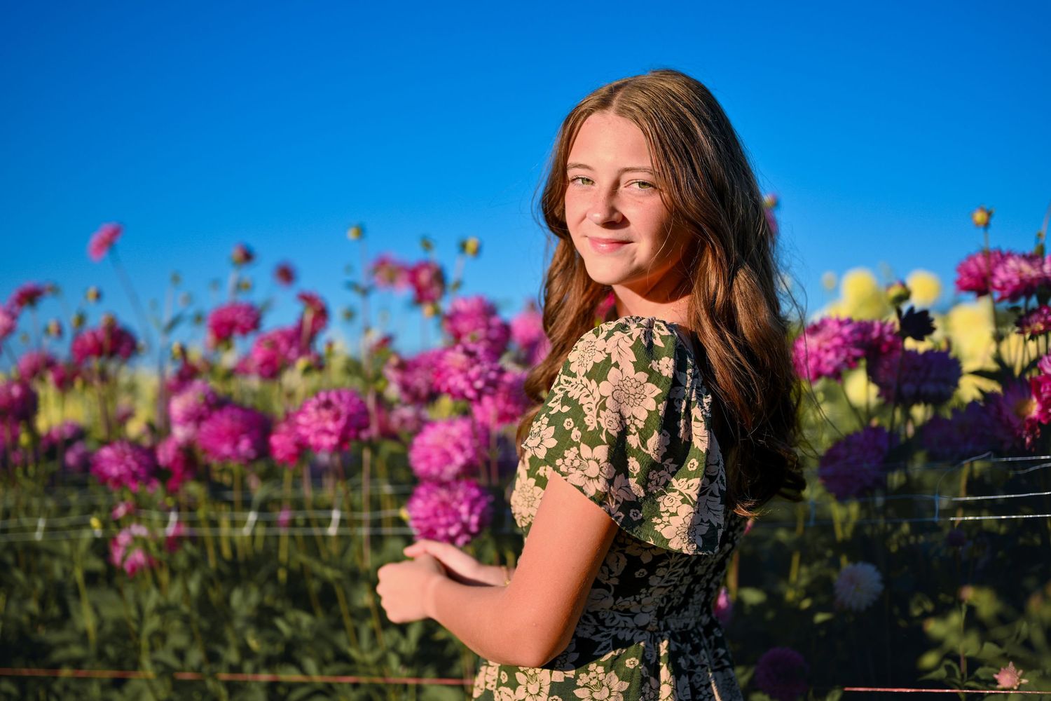 Young girl in floral dress smiling among vibrant pink dahlia flowers under a clear blue sky.