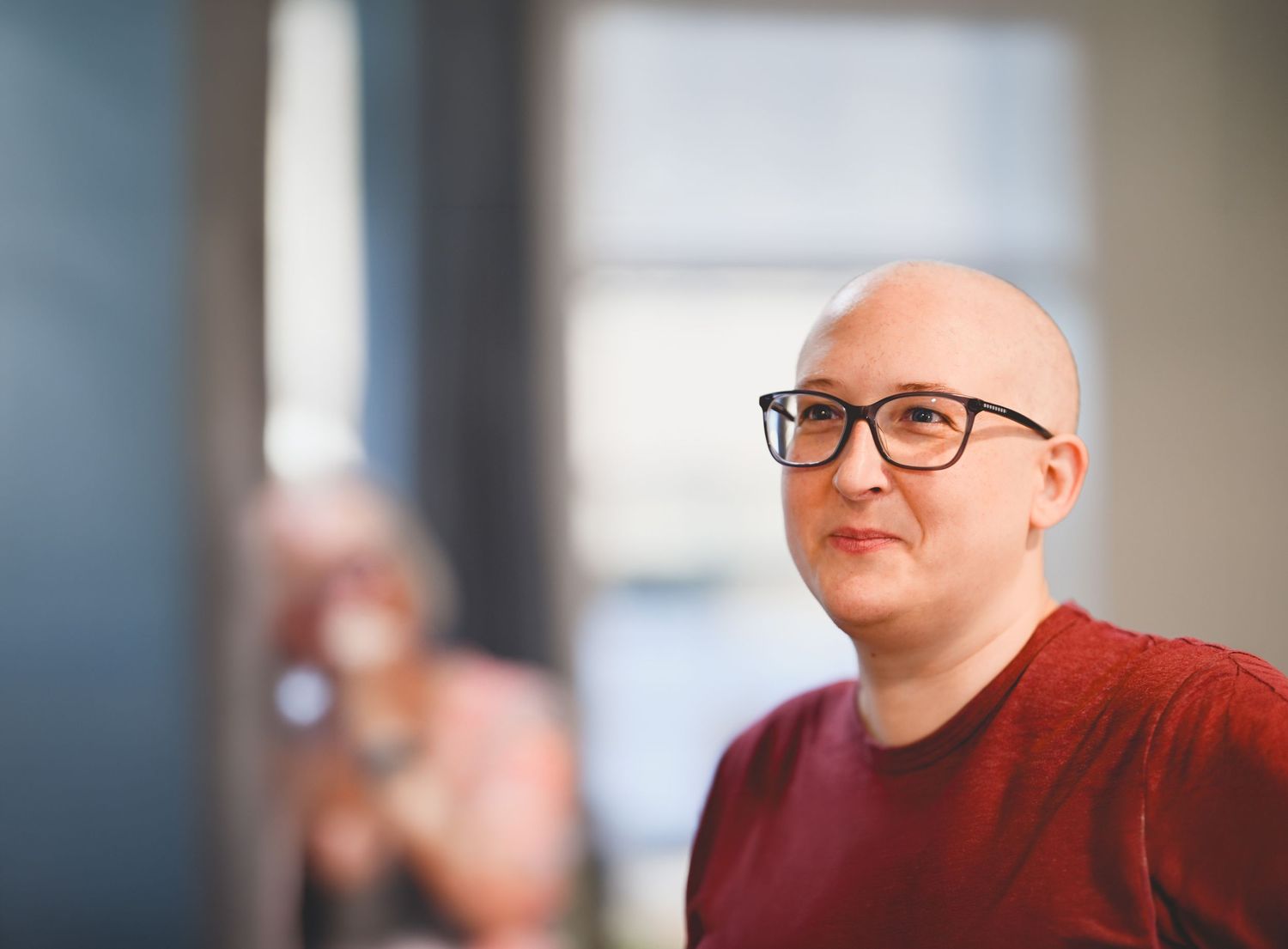 Bald person wearing black glasses and red shirt in a modern office setting.