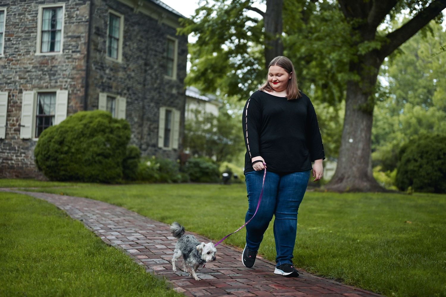 Woman in black top walking small gray dog on brick path beside stone building and green trees.