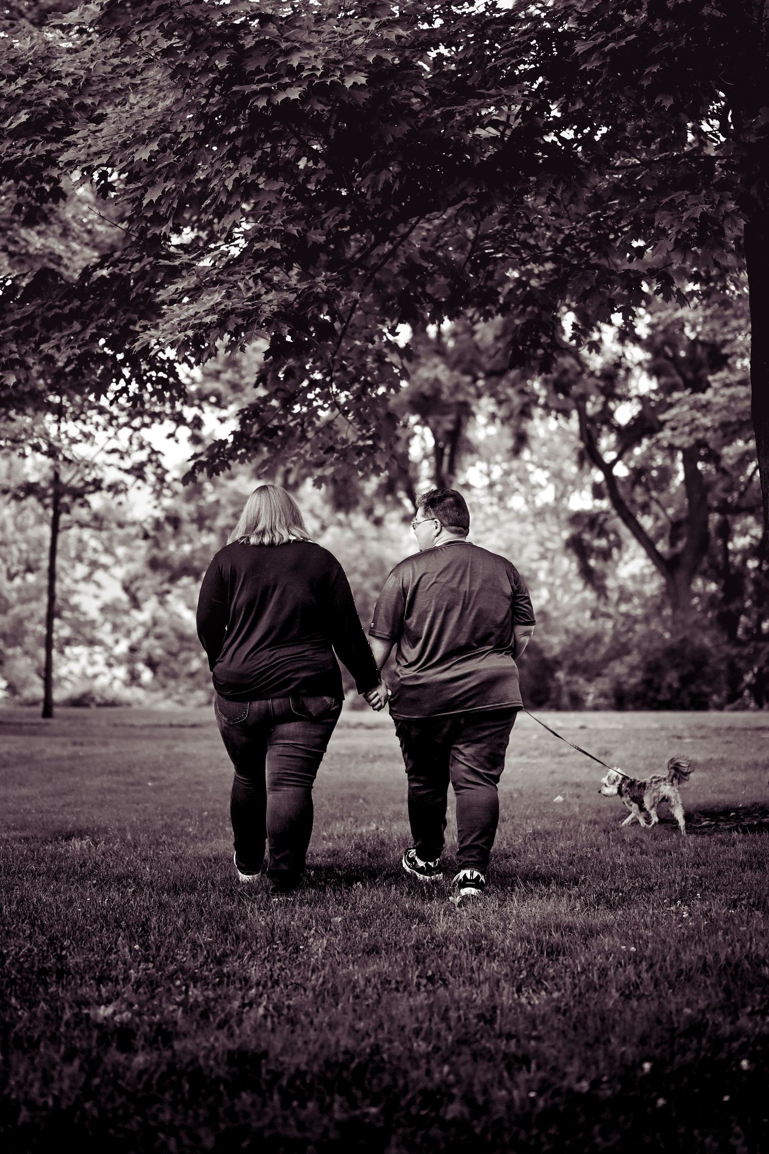 A black and white photo of a couple walking their small dog together on a grassy path beneath leafy trees.