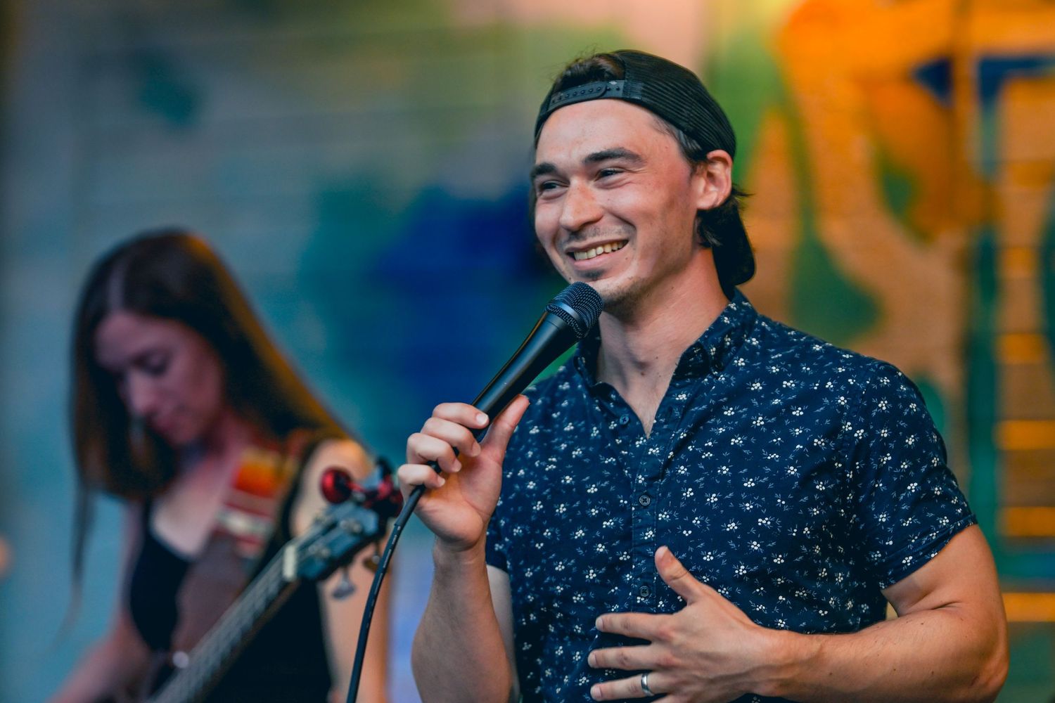The Mimic band: A person in a navy polka dot shirt performs on stage with a microphone while smiling at the audience.
