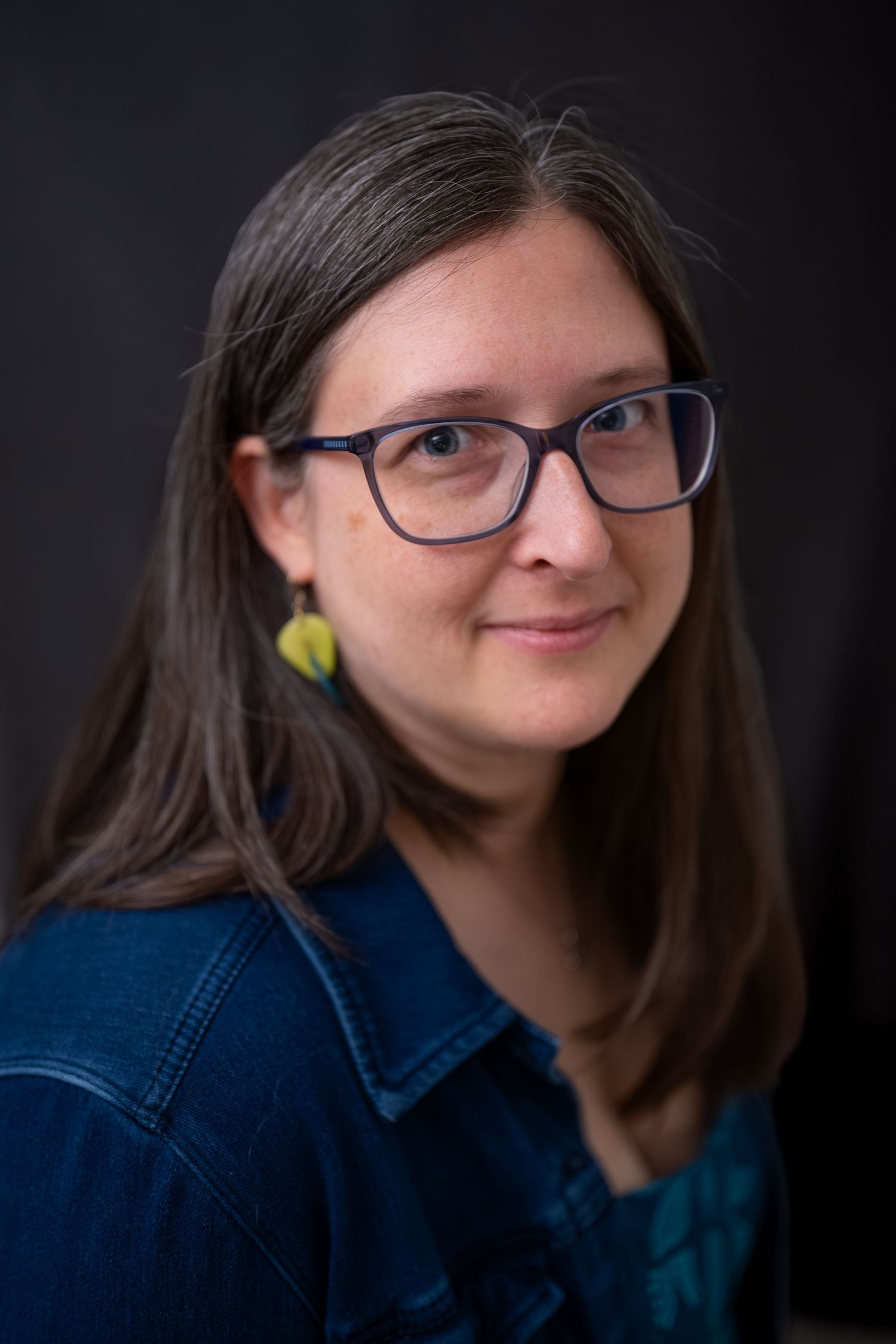 Woman with long brown hair, blue glasses, and yellow earrings wearing a denim jacket, smiling in a portrait photo.