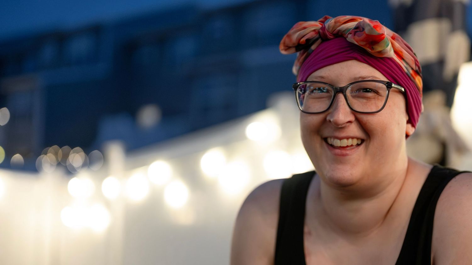 Smiling woman wearing colorful headscarf and glasses outdoors at dusk with bokeh string lights in background.