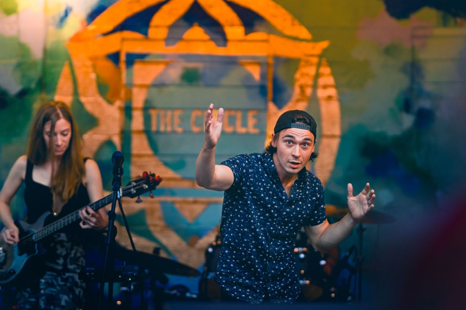 Live band performs on stage with a musician in front of an orange geometric backdrop during a concert.