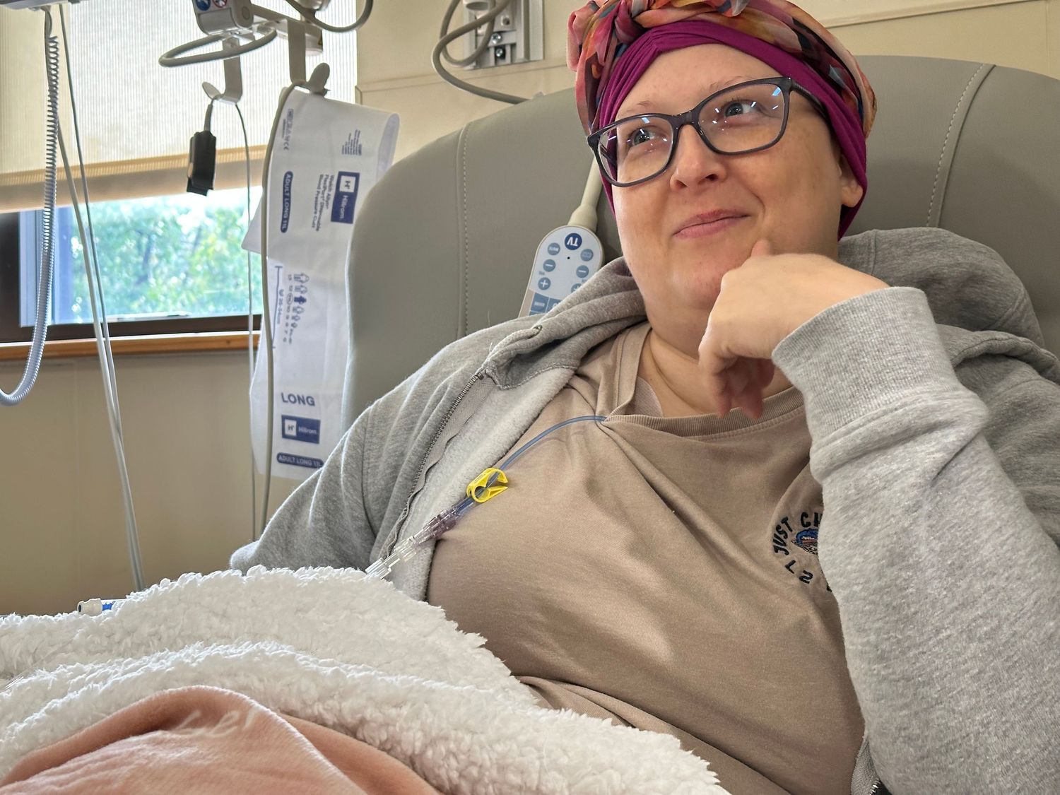 Woman in headscarf and glasses resting in hospital bed, wrapped in blanket, looking calm and resilient.