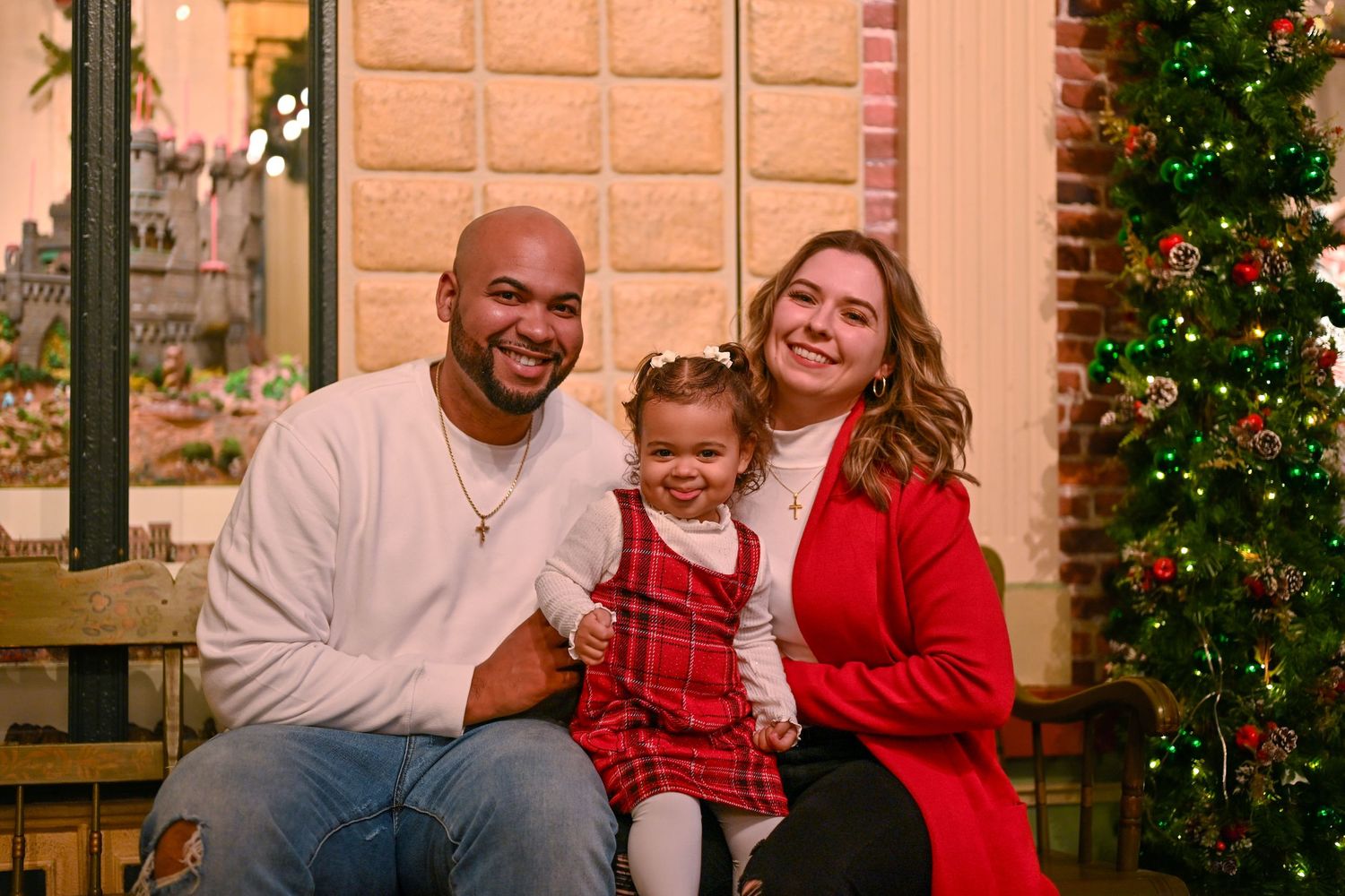 Family of three poses by Christmas tree, parents in red and white holding toddler girl in plaid dress.