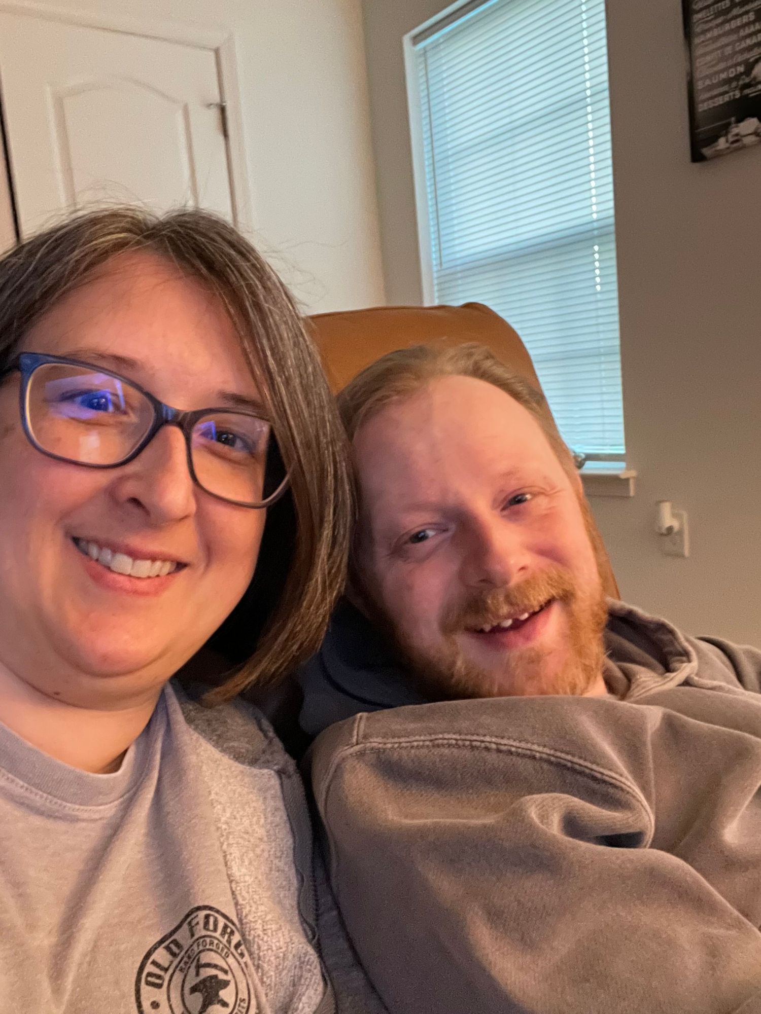 A smiling woman with glasses and a man with a red beard take a cozy selfie together indoors.