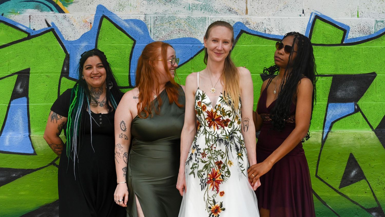 People gathered outdoors on a sunny day with colorful graffiti art on a wall in the background.