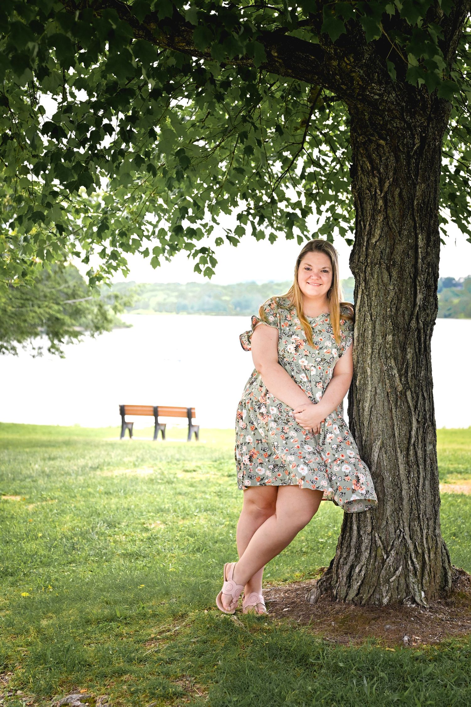 Summer portrait of a woman in a floral dress leaning against a large tree near a lake with benches in background.