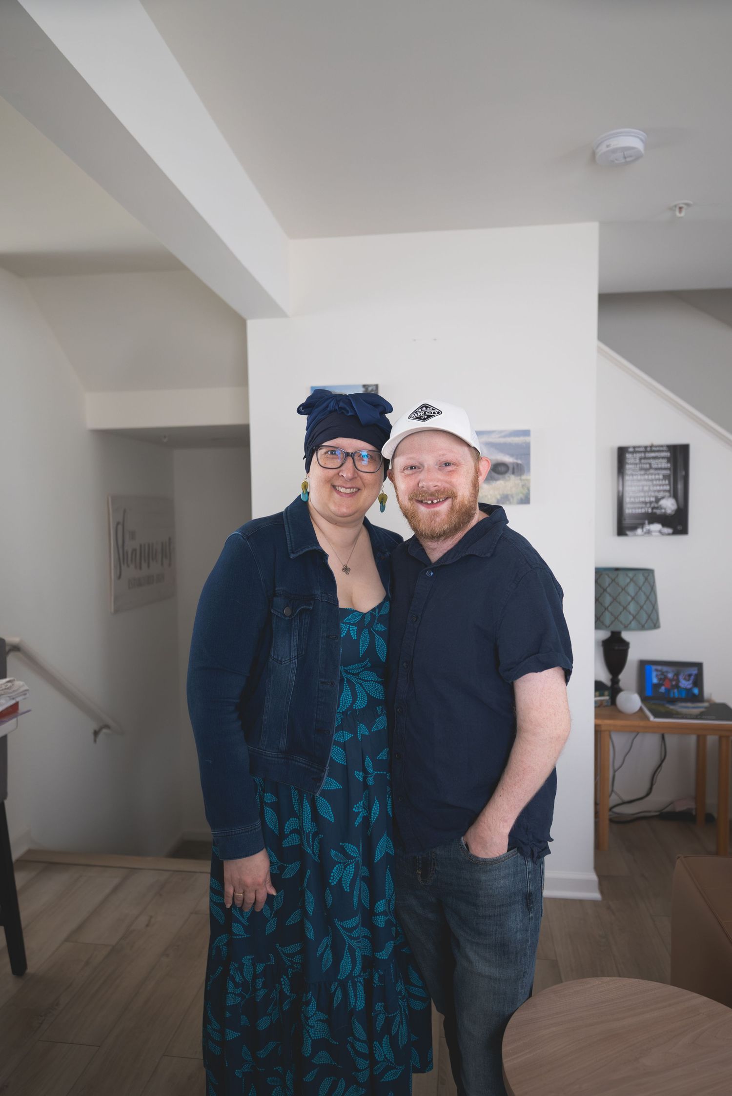 A smiling couple poses together indoors, woman in floral dress with head wrap, man in navy polo and white cap.