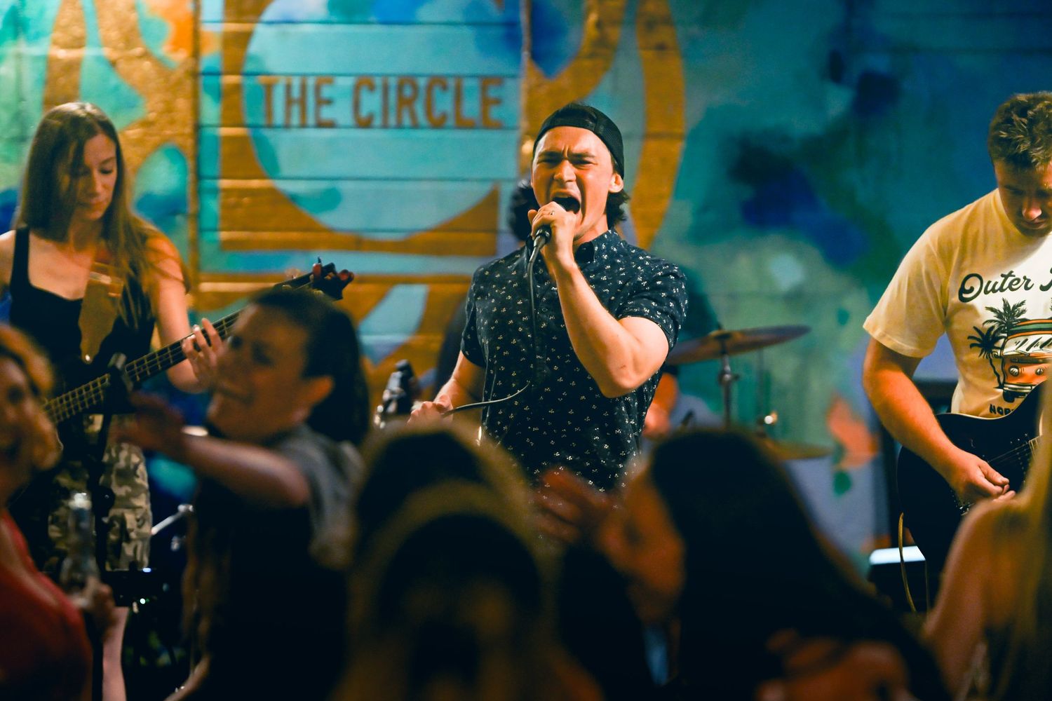 Band performing live at The Circle venue, lead singer with microphone, guitarist visible, energetic crowd in foreground.