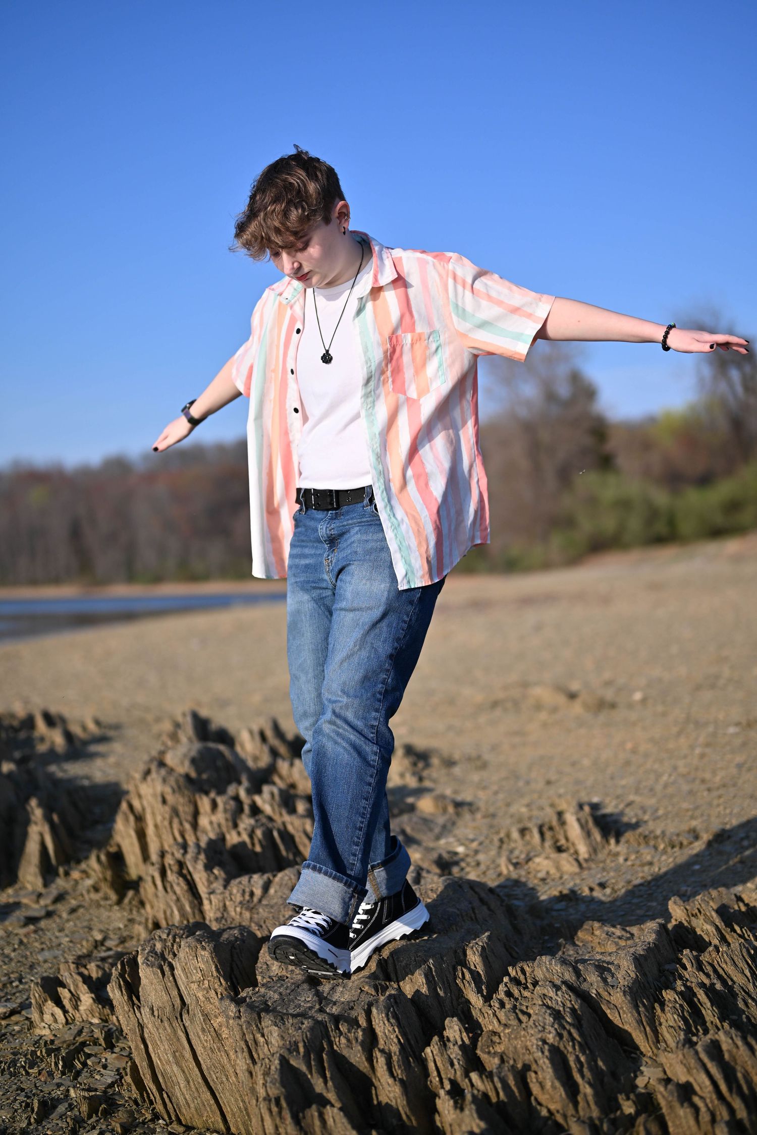 Teen balancing on rocks at sandy beach, wearing striped shirt, jeans, and Converse sneakers on sunny day.