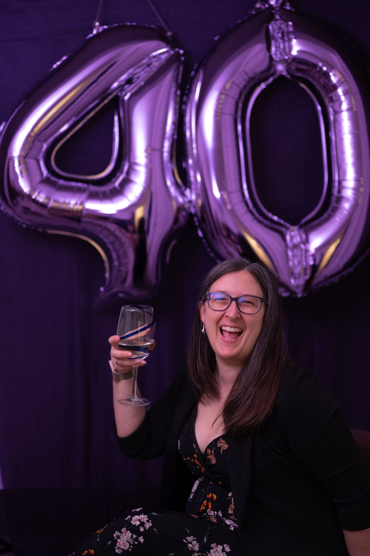 Woman laughing and raising a wine glass in front of purple 40th birthday balloons at a celebration party.