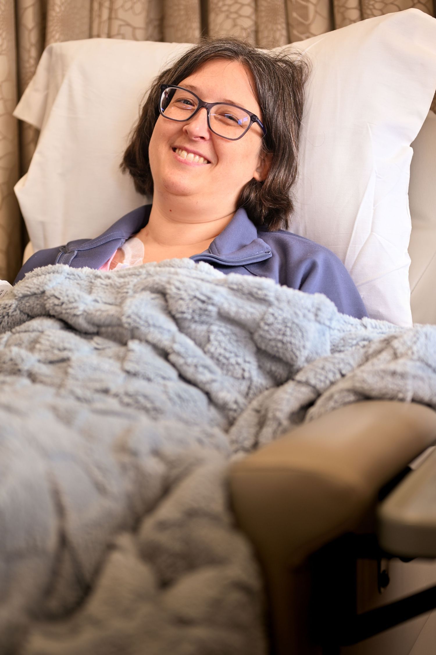 Smiling woman with glasses lying in hospital bed covered with soft gray blanket, looking relaxed and cheerful.