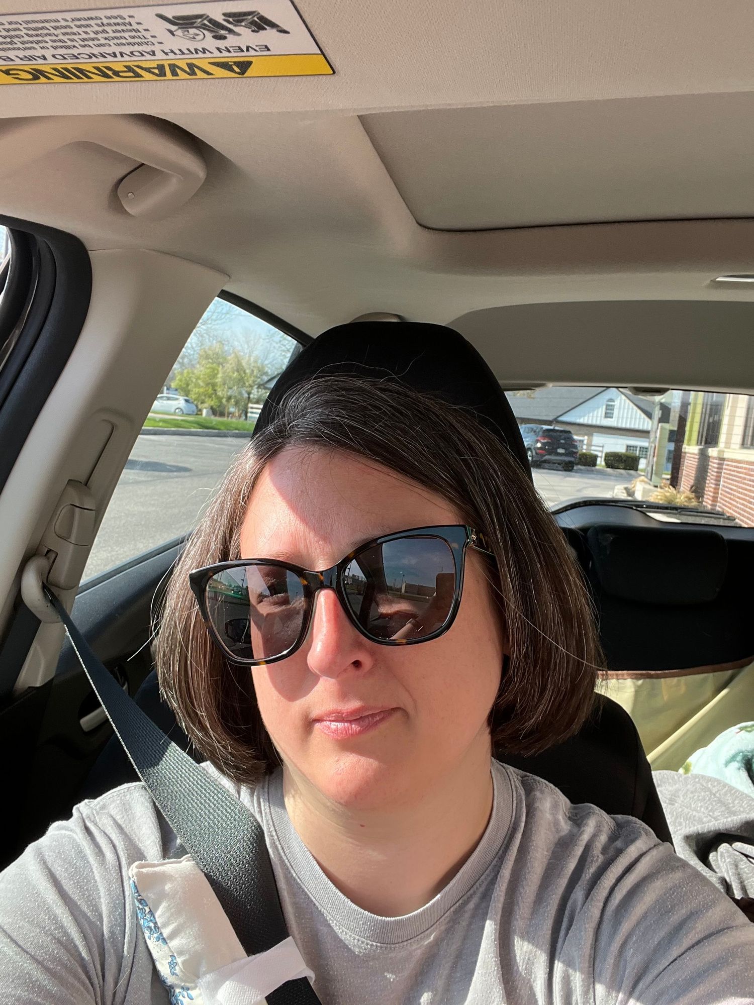 Woman wearing sunglasses taking a selfie inside a car on a sunny day in a parking lot.