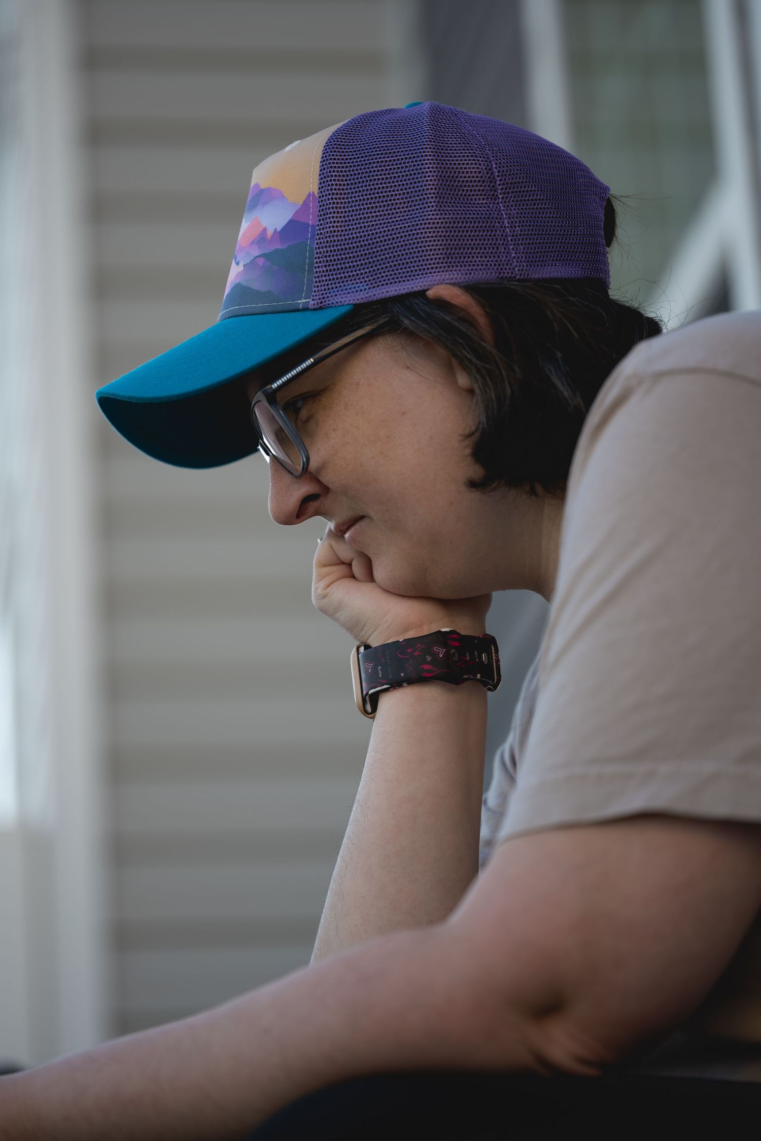 A smiling young person wearing a purple and blue trucker hat and glasses, resting chin on hand outdoors.