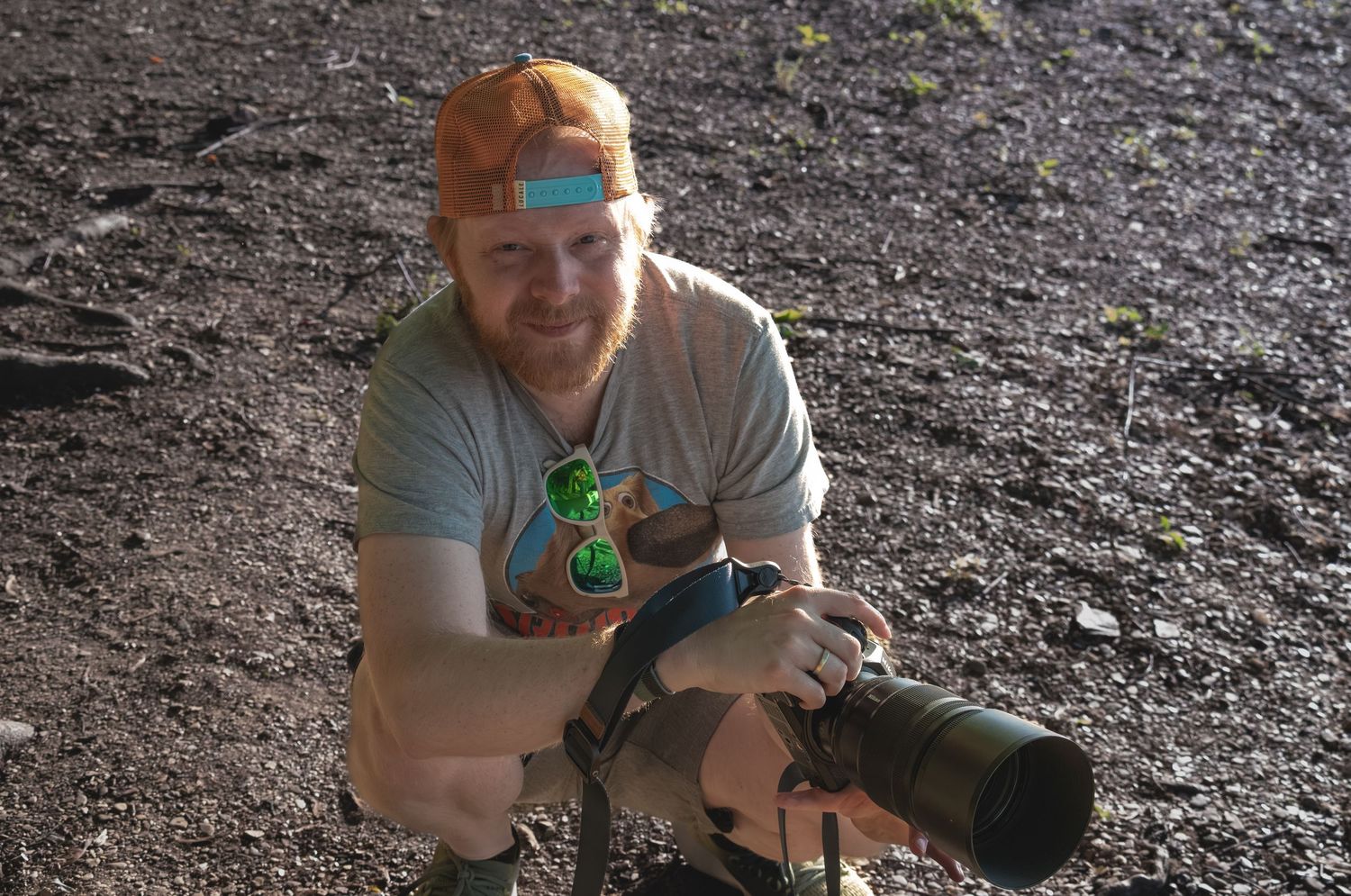 Man crouching outdoors holding a large camera lens, wearing an orange cap and casual t-shirt on rocky terrain.