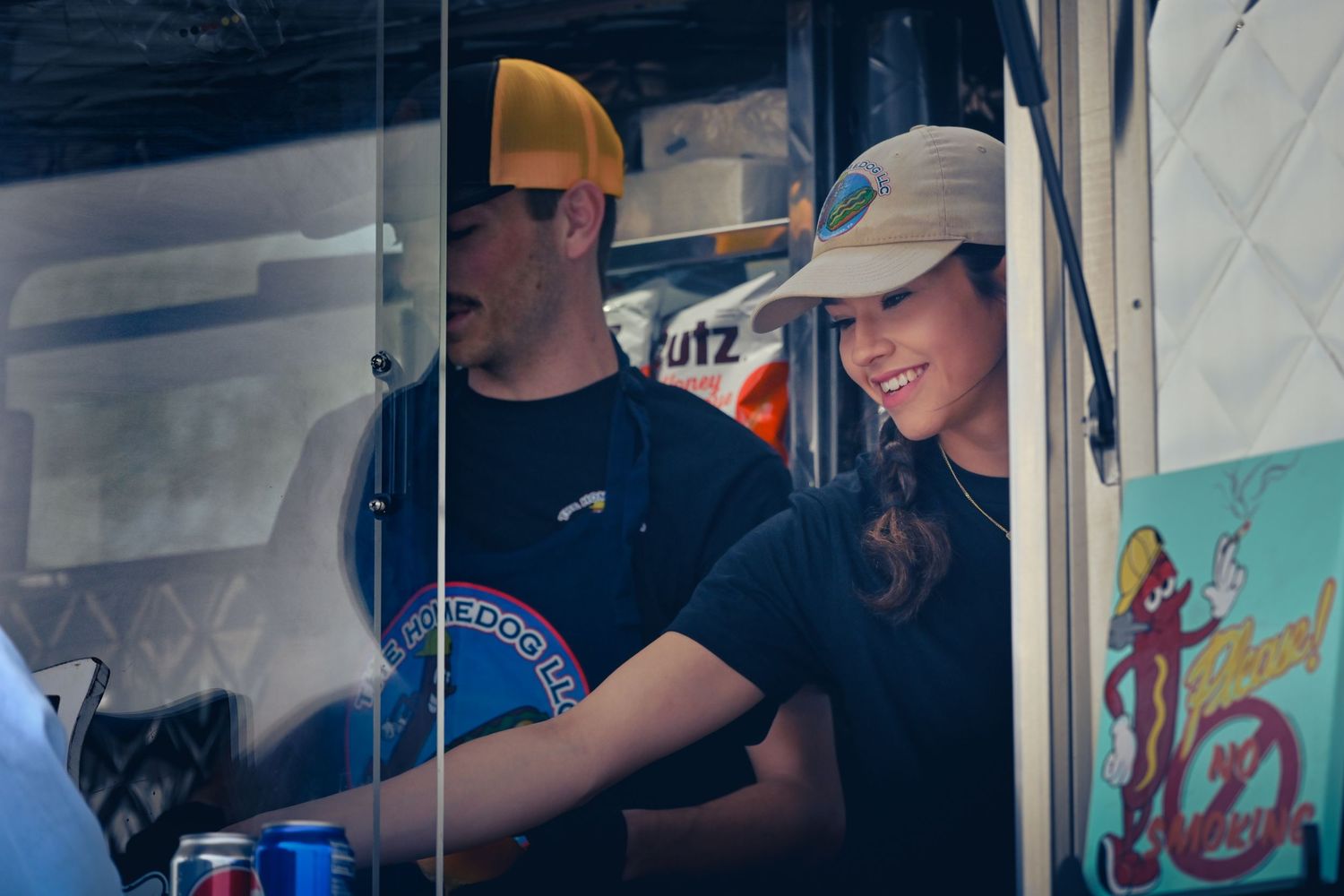 Two food truck workers serve customers from inside their truck window on a sunny day.