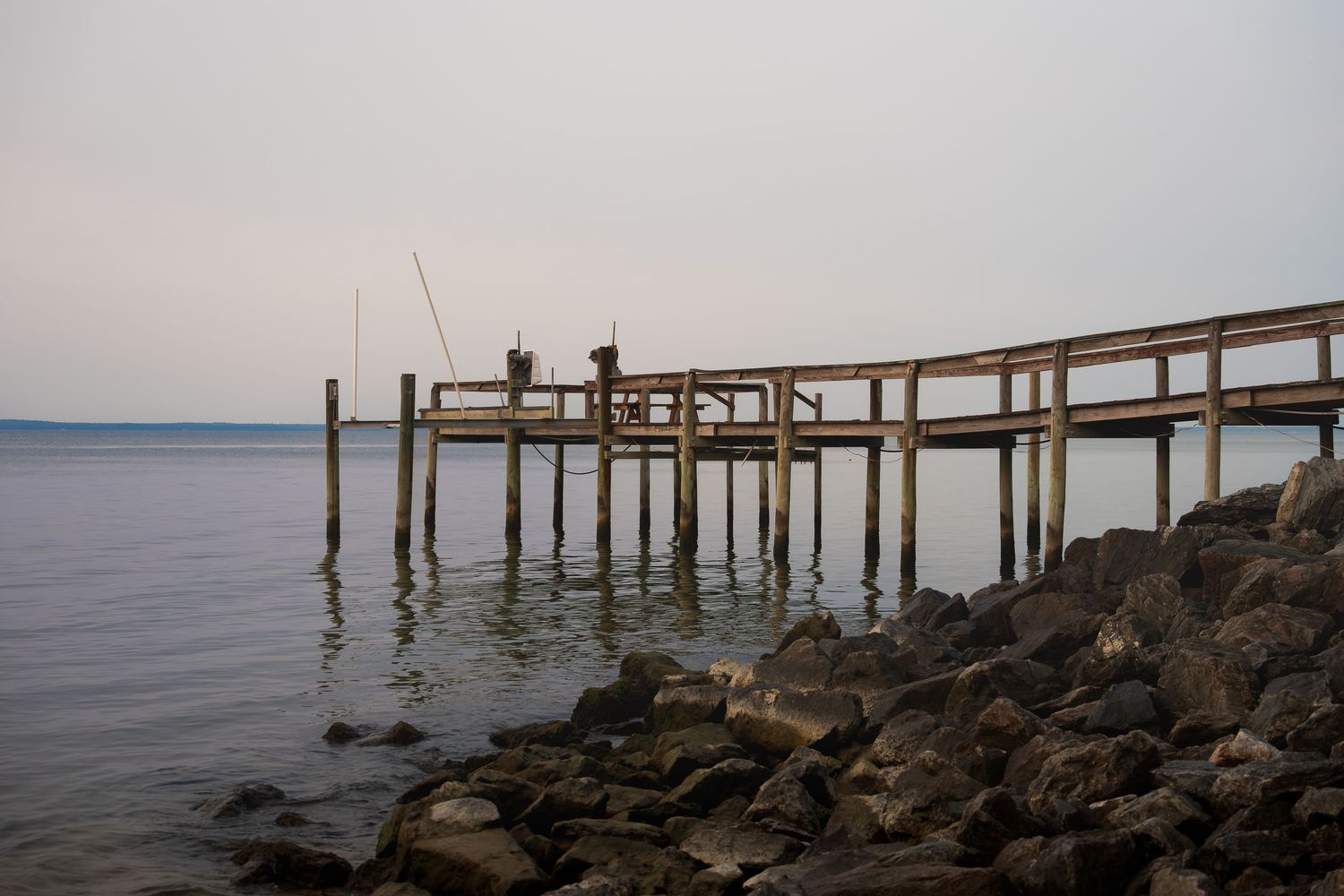 Fishermen stand on a weathered wooden pier over calm water at dusk, with rocky shoreline in the foreground.