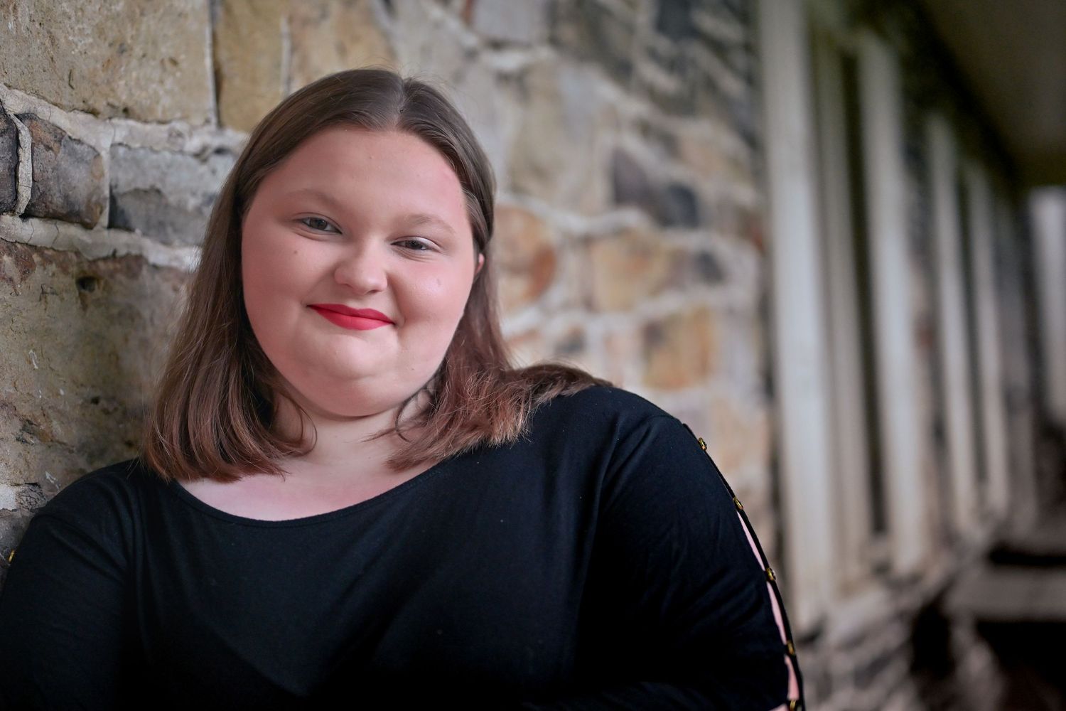 Young woman with red lipstick smiling warmly, leaning against a stone wall outdoors.